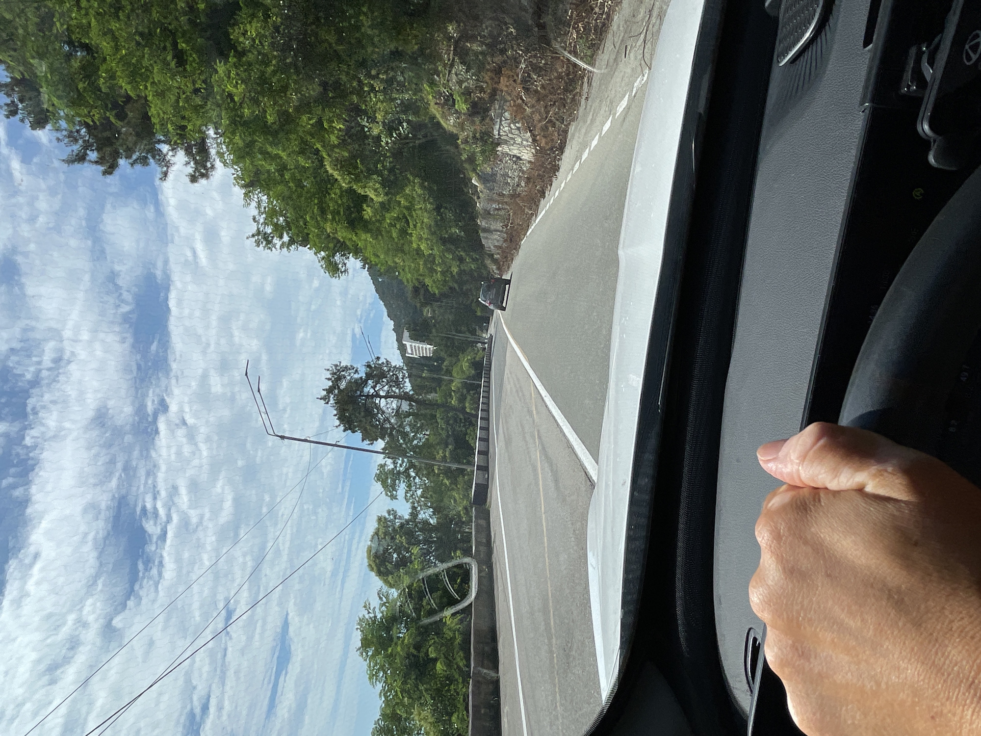 A first-person perspective shot captures a hand gripping a steering wheel as the vehicle travels along a paved road with white lane markings. The road curves to the right, bordered by a concrete barrier on the left and a vegetated embankment on the right, with scattered debris. In the distance, a dark-colored car is visible ahead on the road. The sky is partly cloudy, with a bright blue expanse dotted with white cumulus clouds. Lush green trees line the roadside and climb the hillsides. Utility poles and wires are present along the route. A hint of a building is visible on a distant hillside. The scene suggests a journey through a scenic, possibly mountainous, area.