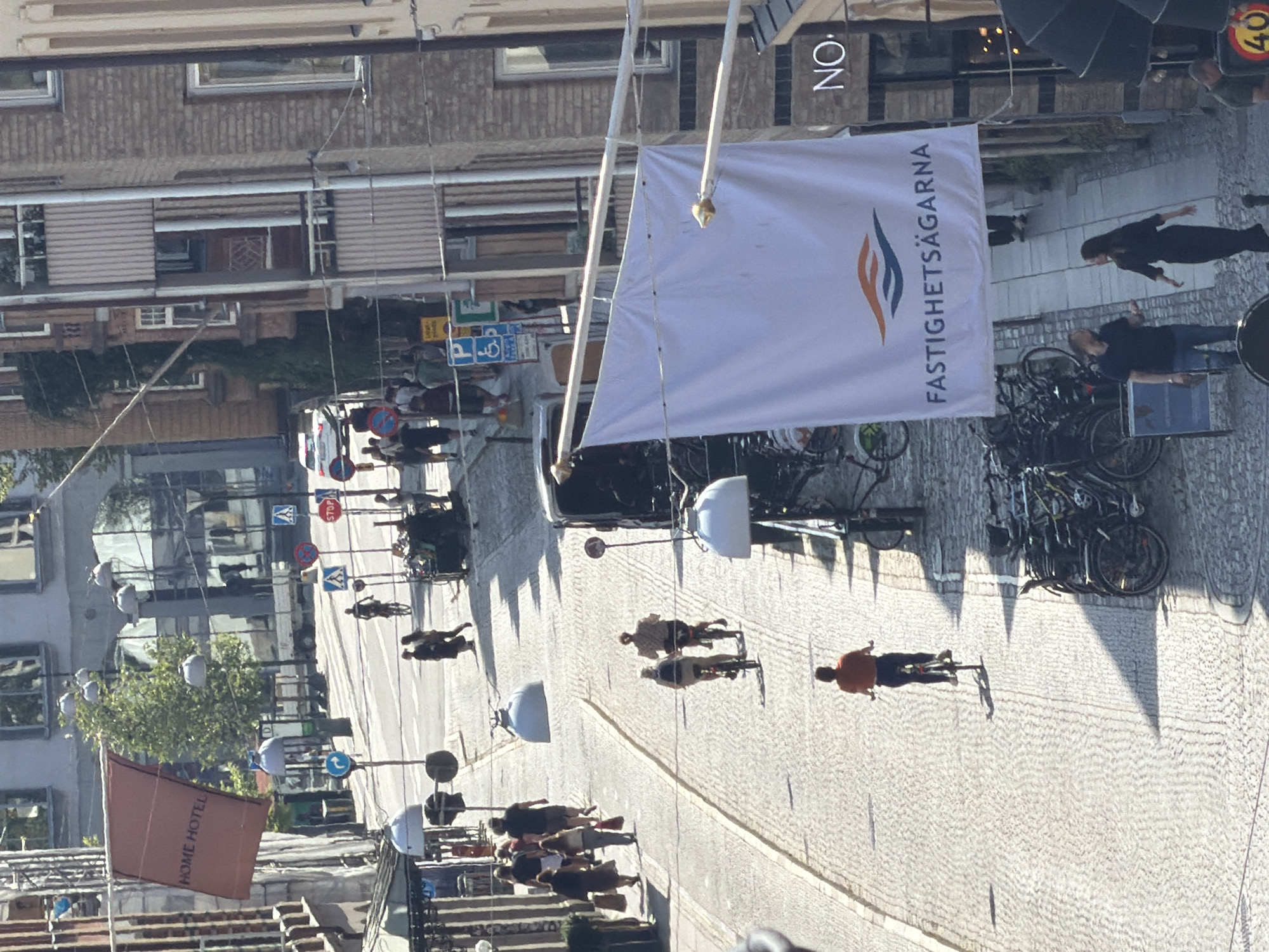 The image captures a street scene in Malmö, Sweden, likely in the vicinity of Gustav Adolfs torg, given the architectural style and the presence of signage. The perspective is from an elevated viewpoint, looking down a cobblestone street lined with buildings of varying architectural periods.
In the foreground, a large white banner with the Swedish text "FASTIGHETSÄGARNA" (Property Owners) and a logo hangs suspended, partially obscuring the buildings behind it. To the right of the banner, a building facade displays the sign "NO." further down the street, the scene unfolds with several individuals and objects.
Several people are seen walking and cycling. Two cyclists are riding away from the viewer, one in a light-colored shirt and the other in a dark top. Further up the street, a single cyclist in an orange shirt is riding towards the viewer. A group of people are walking together on the left side of the street, some carrying bags. Another group of cyclists is parked to the right of the frame.
Various traffic and directional signs are visible along the street, including a blue sign with a white "P" for parking and a wheelchair symbol, and a red circular sign with a white horizontal bar, indicating "no entry" or a similar restriction. Cars and a truck are also present in the background. The street itself is paved with cobblestones, and shadows are cast across the scene, indicating it is a sunny day. The architecture includes buildings with balconies, windows of different sizes, and brickwork, suggesting a mix of historical and more modern constructions.