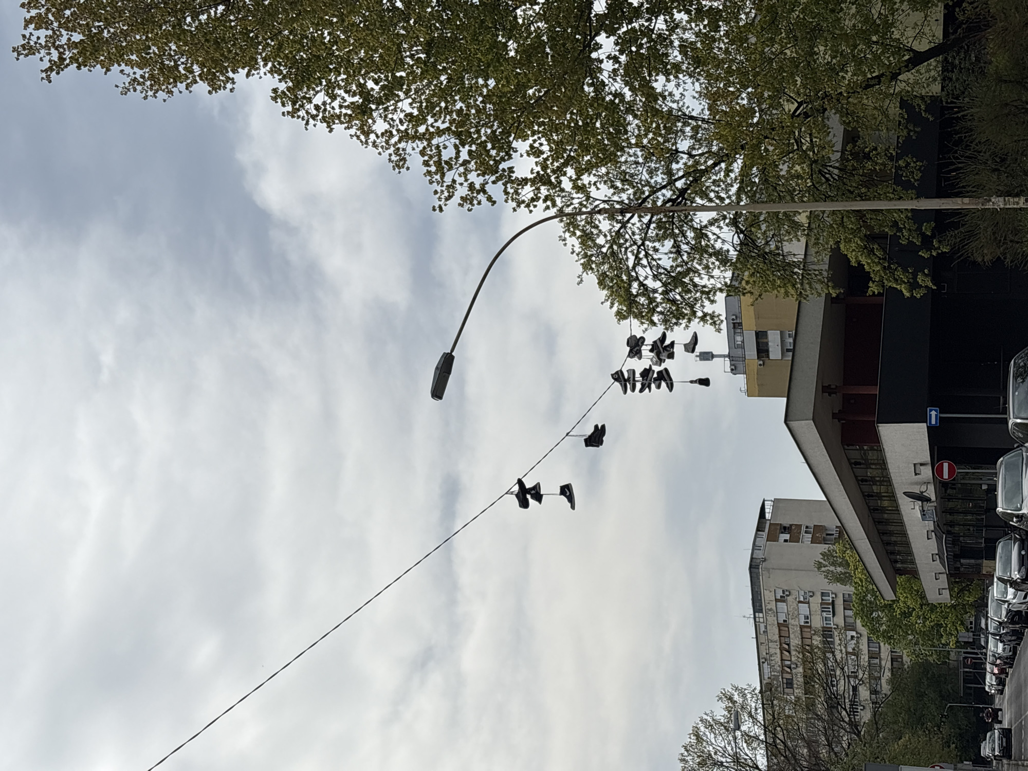 The image shows an urban scene, likely in Belgrade, Serbia, based on architectural styles and street signs. A power line stretches diagonally across the frame, suspended between a street lamp and a building. Several pairs of shoes are hanging from the power line. Buildings are visible in the background, with architectural characteristics common in Belgrade. Cars are parked on the street to the right. The sky is overcast. Tree foliage is visible in the upper portion of the image.