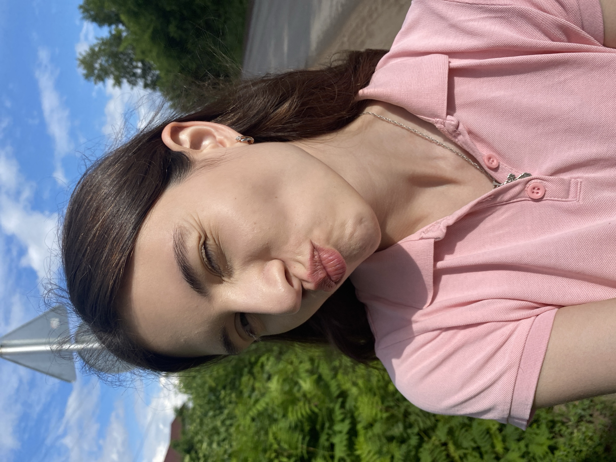A young woman with long, dark hair is captured in a close-up, selfie-style shot outdoors. She is wearing a pink polo shirt and a silver necklace. Her right eye is partially closed in a wink, and her lips are pursed in a playful pout. The background consists of lush green foliage on the left, a paved road and trees on the right, and a bright blue sky with scattered clouds above. A white triangular sign is visible in the upper left portion of the frame. The lighting suggests it is daytime, likely sunny. The scene appears to be in a rural or suburban area, possibly on the outskirts of Votkinsk, Russia, given the visual cues of vegetation, a road, and what could be a residential building in the far left background.