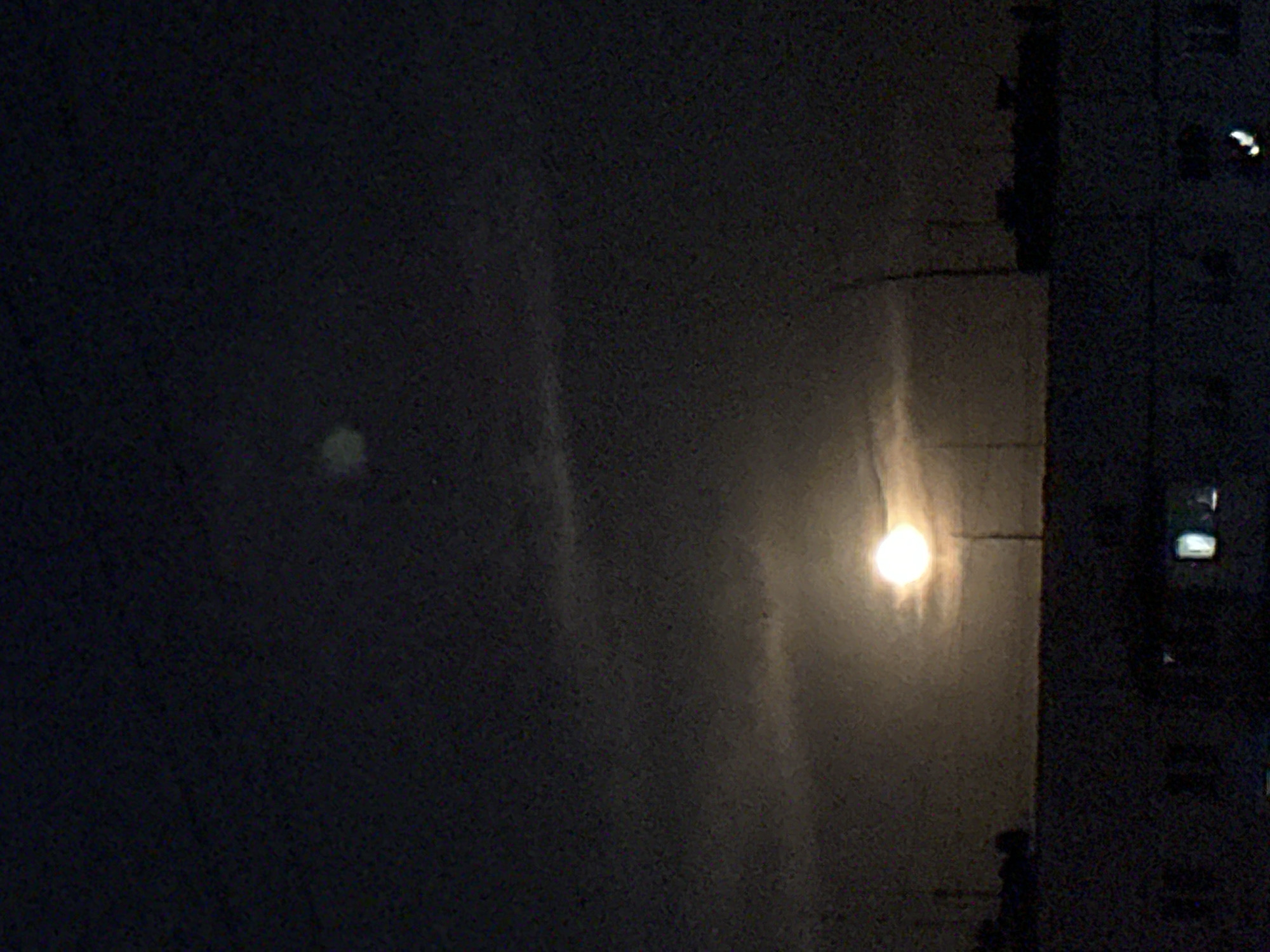 The image captures a nocturnal scene characterized by two luminous sources against a dark, grainy background. On the right side, a prominent, intensely bright, circular light emits a strong, diffused glow with noticeable vertical streaking, suggestive of a full moon or a powerful artificial light viewed through atmospheric conditions such as clouds, fog, or a semi-transparent surface like a dirty window. To the left, a second, significantly fainter and more diffuse circular light is visible.
A dark, architectural structure, likely a section of a building, runs vertically along the far right edge of the frame. This structure exhibits small, rectangular points of light, possibly illuminated windows or reflective surfaces. The overall ambiance is dark and hazy, with a textured appearance indicative of low-light photography. There are no discernible people or activities, and no text is visible. The scene portrays a tranquil, late-night urban or suburban environment.