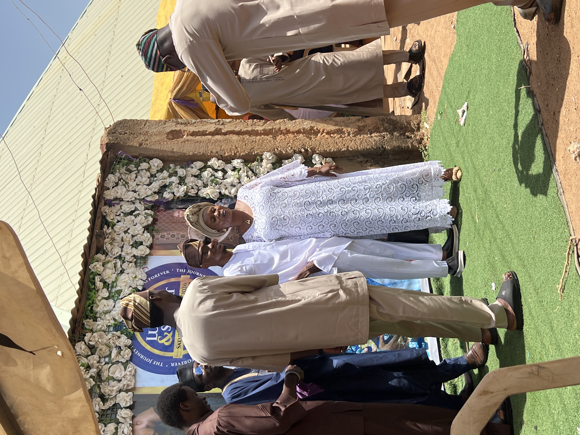 The image depicts a group of people standing outdoors in what appears to be Rigasa, Nigeria. There are at least seven individuals visible, all of whom appear to be African. Most of the men are wearing traditional Northern Nigerian attire, while a woman is wearing a white lace gown and headwrap. They stand in front of a memorial backdrop, adorned with white flowers and a poster bearing a photograph. The ground is covered in part by green artificial turf and sandy soil. The scene suggests a funeral or memorial service taking place in an open space.