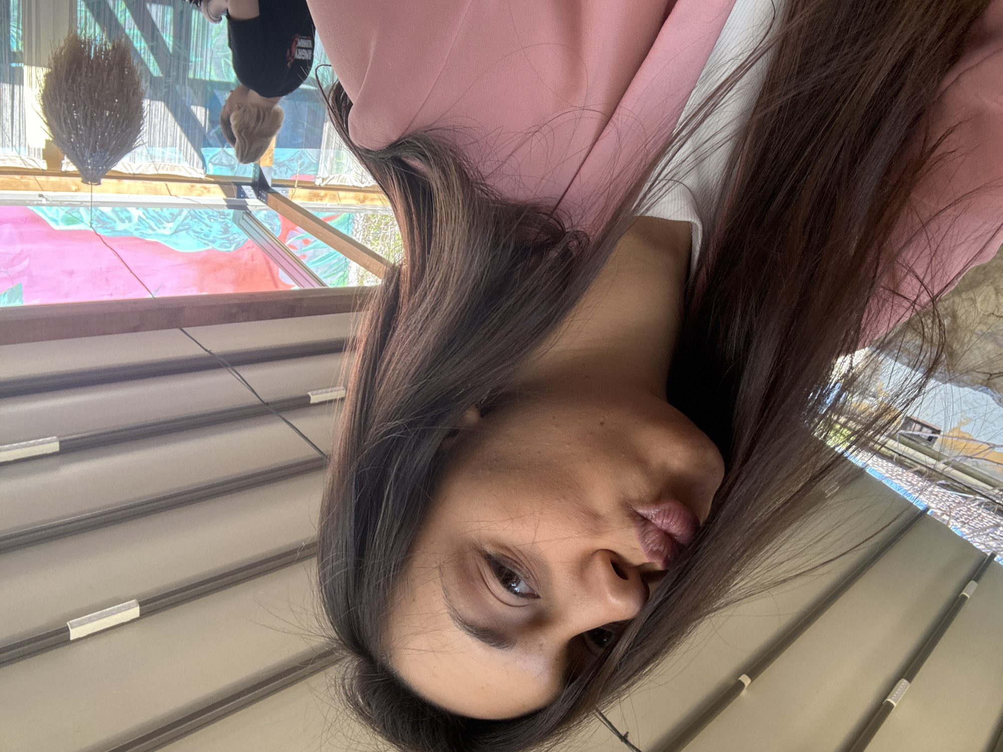 The image shows a woman with long, brown hair, wearing a pink blazer over a white top. She is looking at the camera with pursed lips. The background includes a ceiling with blinds and a window reflecting a person and a building with a decorative object hanging from the ceiling. The location is Kragujevac, Serbia, based on the background details.