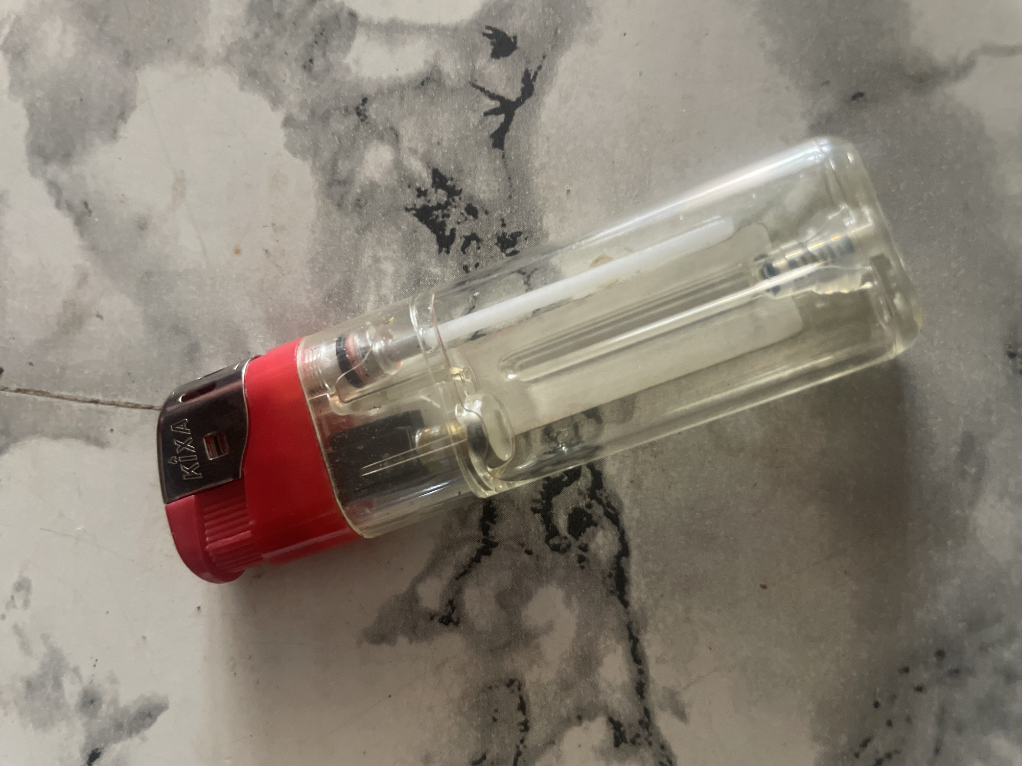 A red and clear disposable lighter is lying on a grey, marbled surface. The lighter has the brand name "KIXA" printed on a silver metal plate at the top. The surface appears to be a floor or countertop with dark grey and black marbled patterns. The scene is lit from above, casting subtle shadows. There are no people or any other objects present. The location of New Stadium, Nigeria is not visible or inferable from the image.