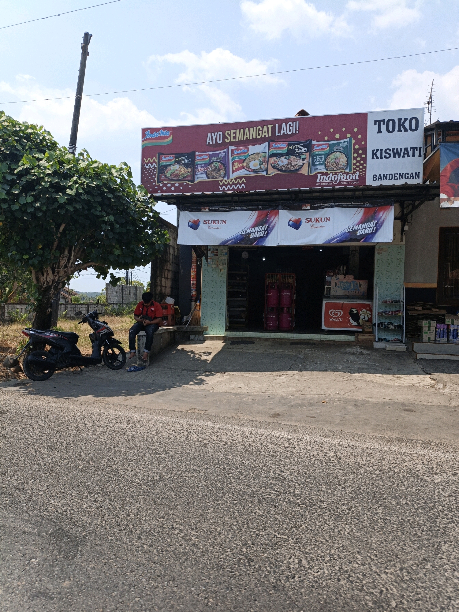 The image depicts a street view in Cikal, Indonesia, featuring a small storefront with a prominent billboard advertising Indofood products. The billboard, with its bold red and white lettering, displays various Indomie instant noodle flavors and a prominent "Indofood QUALITY FOODS" logo. Below the billboard, a banner reading "SUKUN Executive SEMANGAT BARU!" is displayed.
The storefront itself appears to be a convenience store or small market, with merchandise visible through its open entrance. Notable items include large pink gas cylinders, a Wall's ice cream freezer, and shelves stocked with various goods, including what appear to be cleaning products or sprays.
In front of the store, a person in a red shirt and dark pants is seated on a concrete ledge, looking at their phone. Two motorcycles are parked on the roadside. A large, leafy tree provides shade over the area. The scene is set against a backdrop of a clear blue sky with scattered clouds, and the overall environment suggests a warm, sunny day. The road surface is asphalt, with some concrete paving near the storefront.