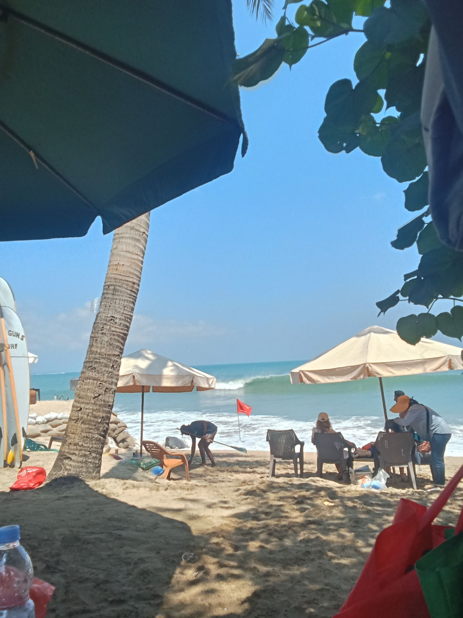 The image depicts a beach scene in Kuta, Indonesia, likely on a sunny day. In the foreground, a dark shaded area suggests the presence of an umbrella or overhang. A clear plastic water bottle is visible on the left side. The sandy beach leads to the ocean where waves are breaking. A palm tree stands prominently on the left, with two surfboards leaning against it. Beach chairs are scattered on the sand. A person is actively raking the sand near a light-colored umbrella. Further back, near another umbrella, two people are seated in chairs, one wearing a hat. Another person wearing an orange hat and blue jeans is standing and appears to be interacting with items on a table or chair. A red flag is planted in the sand near the water. In the background, the bright blue sky meets the ocean.