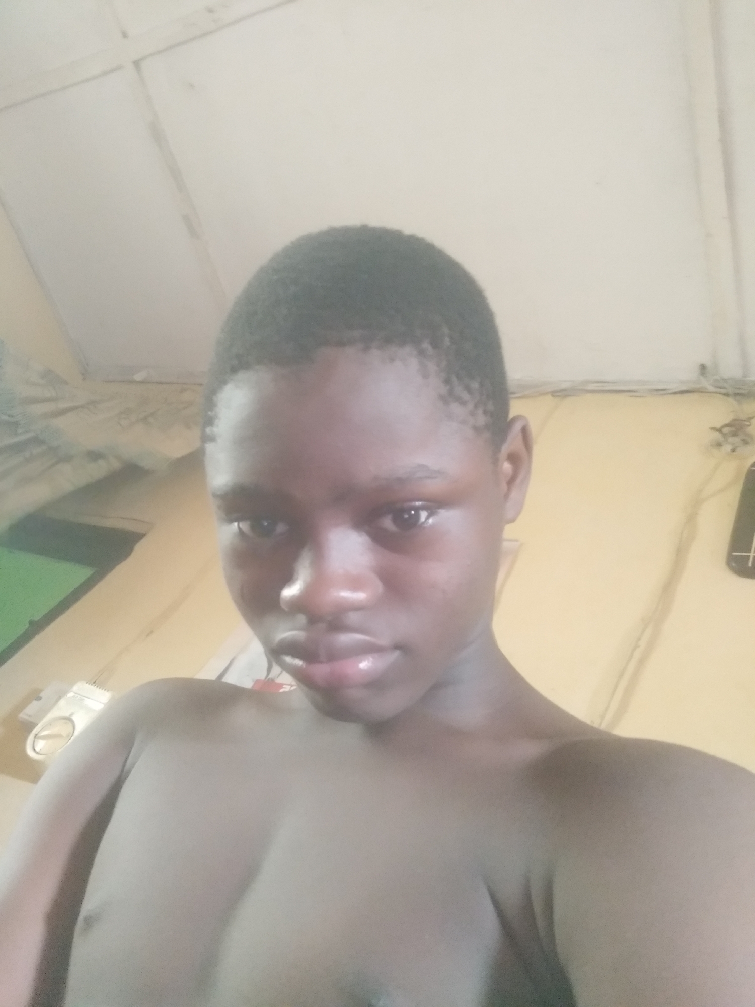 A close-up, selfie-style image shows a shirtless young Black person with short dark hair. The background consists of a white ceiling and a yellow wall with some objects or fixtures attached. The location is Unknown, Unknown.