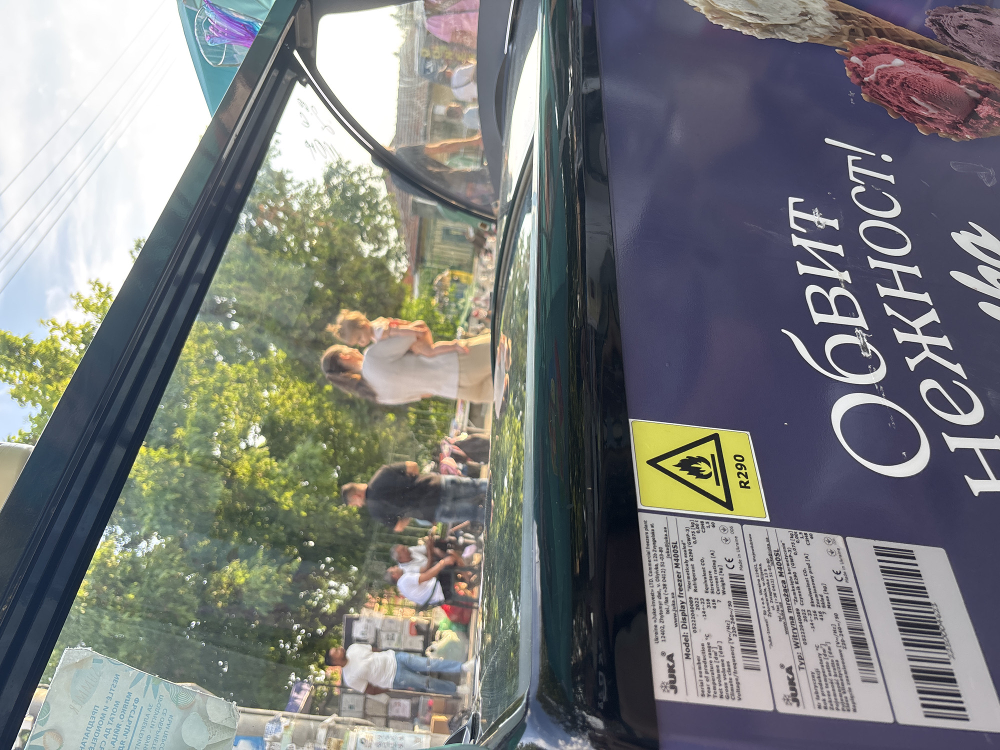 The image depicts an outdoor scene in Kostievo, Bulgaria, viewed through the transparent top panel of a dark-colored JUKA M400SL commercial display freezer. The freezer's label identifies its Ukrainian manufacturer as "Juka-invest" LTD and specifies R290 refrigerant with a corresponding flame warning symbol. Bulgarian branding text "Обвит нежност!" (Wrapped in tenderness!) is visible on the freezer's side, alongside images of ice cream cones.
The scene beyond the freezer's glass, appearing somewhat refracted, shows a group of approximately seven individuals in what appears to be a public or market area during late afternoon or early evening, indicated by warm sunlight. From left to right: a man in a white t-shirt and blue jeans stands beside stacked containers; two men are seated on the ground; a woman in light clothing holds a young child; and a man in a dark t-shirt stands facing away. Another partial figure is visible on the far right. Various bags and items are scattered on the ground near the seated individuals. The background features lush green trees, partially obscured buildings with tiled roofs, and a clear blue sky with overhead power lines. A utility pole adorned with numerous Cyrillic-script posters is visible on the far left.