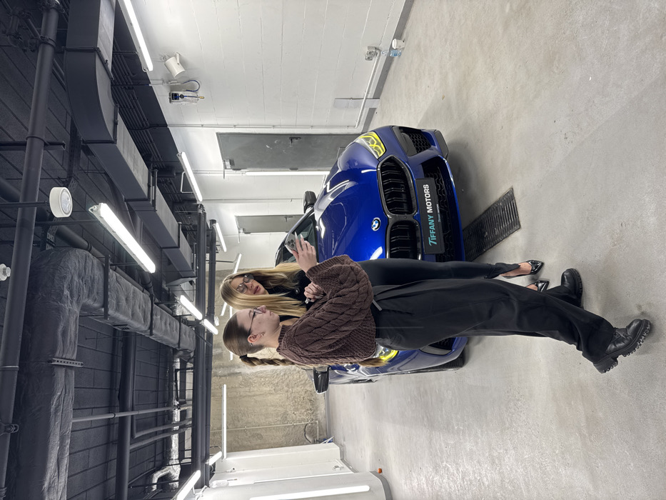 Two women are standing in front of a blue car. The woman on the left is wearing a brown sweater and black pants, and her hair is in pigtails. She's looking at a phone. The other woman has long blonde hair and is wearing black pants and a black top. The car is labeled "TIFFANY MOTORS". The setting looks like a brightly lit garage or a detailing shop with visible ceiling infrastructure, lights, and a drain.