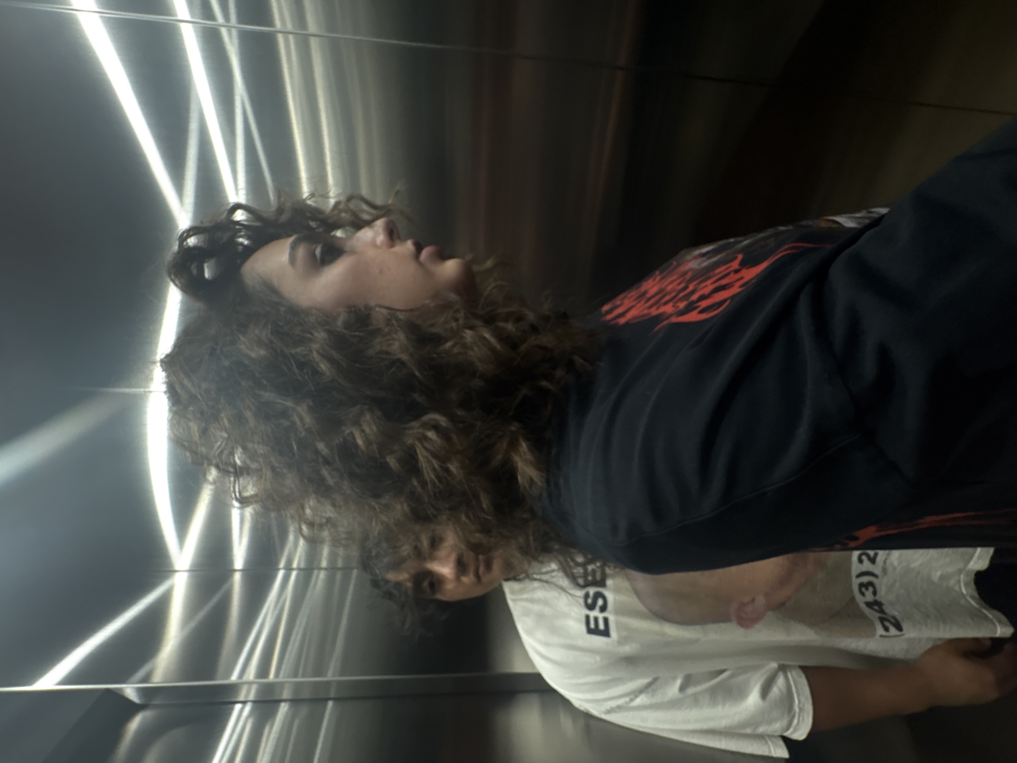 Two people are inside an elevator. One individual, with curly brown hair, is wearing a dark long-sleeved shirt with red flames. The second person, partially obscured, is wearing a white t-shirt with text on it. The elevator has a metallic interior with a crisscross lighting pattern. The location is Mexico City, Mexico.