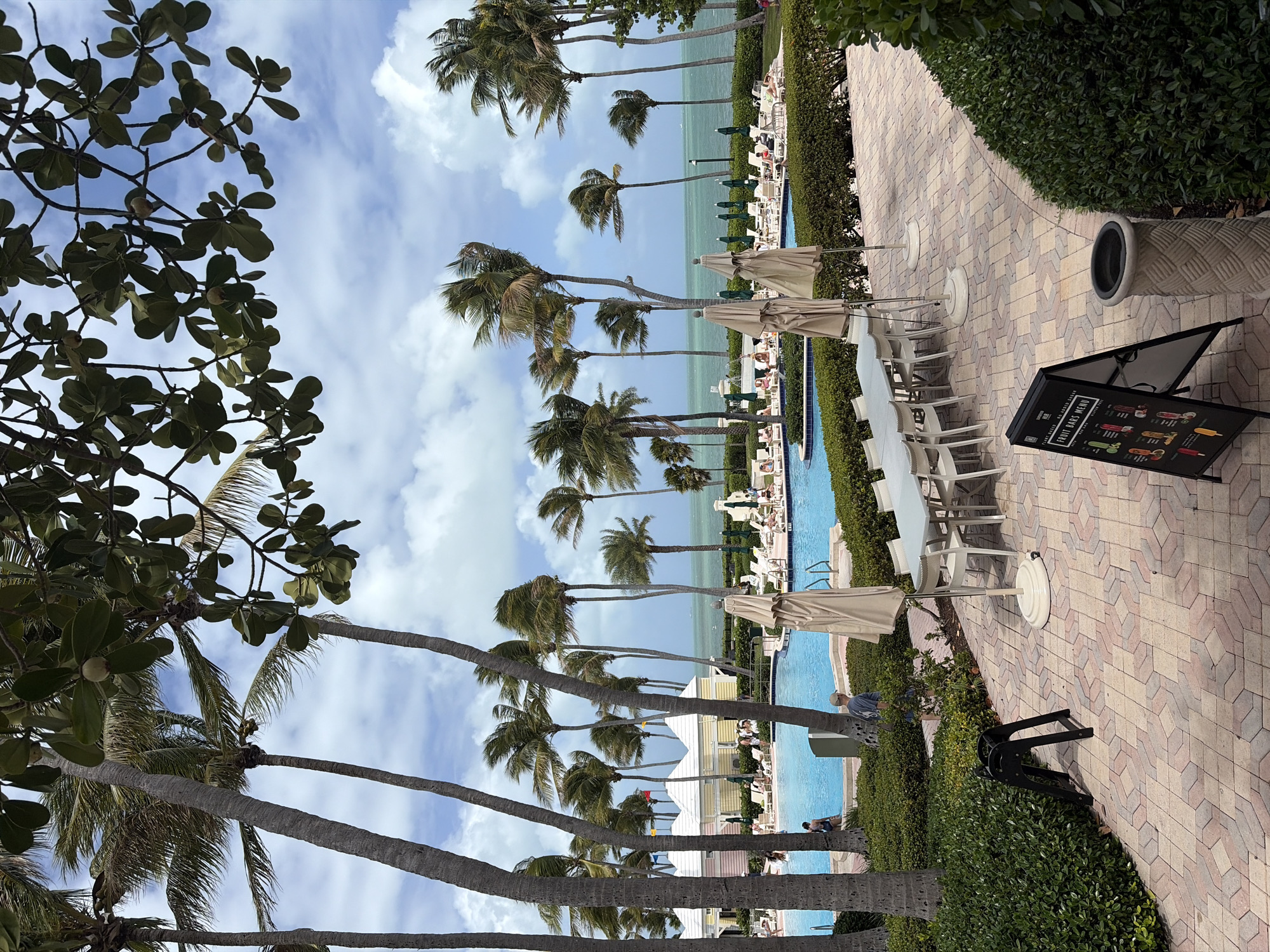 The image depicts a scene in Key Biscayne, United States of America. It features a pool area with palm trees lining the waterfront. Tables and chairs with closed umbrellas are arranged along a brick-paved path, bordered by manicured hedges. In the foreground, there's a tree with lush green leaves. The pool is flanked by lounge chairs and umbrellas, occupied by people. A standing menu board is visible on the brick path. The sky is blue with scattered clouds.
