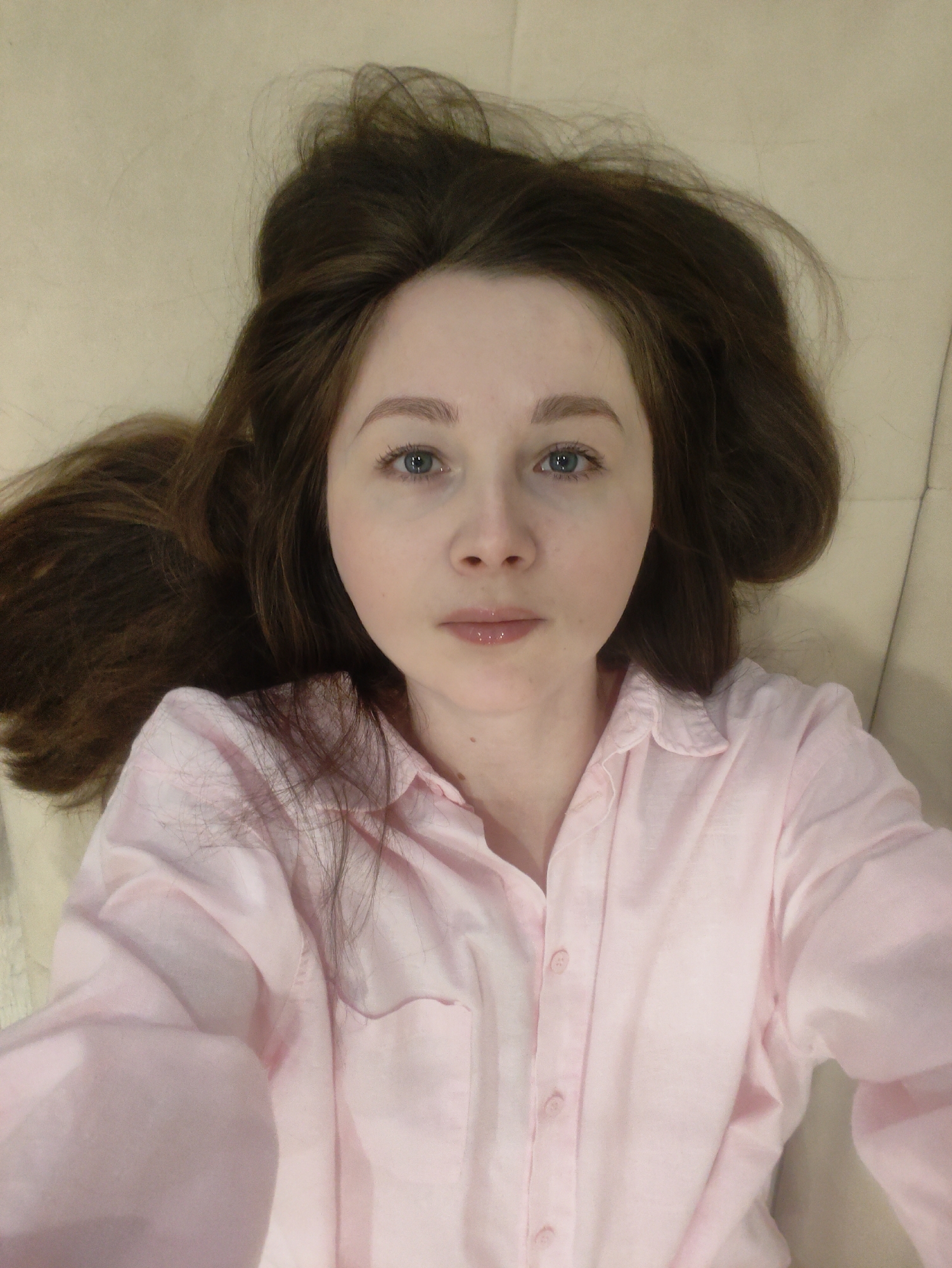 A fair-skinned woman with blue eyes and light brown hair is lying on a light-colored surface, possibly a couch or bed. Her hair is spread around her head. She is wearing a light pink button-down shirt. The location is Unknown, Unknown.