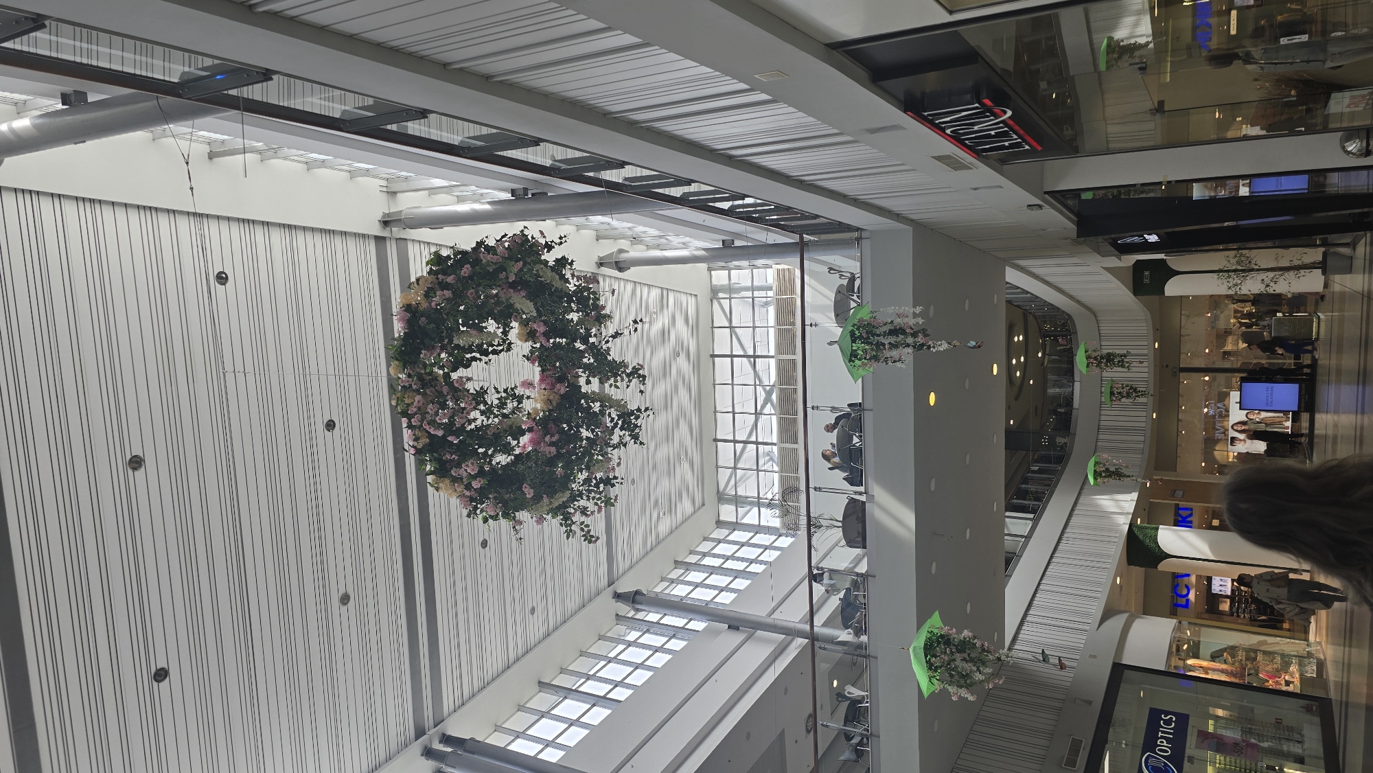This image depicts the multi-level interior of Burgas Plaza Mall in Burgas, Bulgaria. The central feature is a large, circular wreath of artificial pink and white flowers and green foliage, suspended by wires from the uppermost ceiling. The ceiling is white with linear metallic accents and recessed lighting.
An upper floor, visible through a glass railing, hosts a seating area resembling a café. Multiple round tables and chairs are occupied by approximately five to six individuals. Potted plants are also present on this level. Large windows provide significant natural light.
Below, multiple retail storefronts are visible on the lower levels. Signs for "LAZZARINI" and "NY OPTICS" are clearly legible, with "LC WAIKIKI" partially visible. Shop windows display merchandise and mannequins. Smaller floral arrangements, composed of white and pink flowers suspended from green umbrella-like structures, hang from the ceiling over the lower levels. A large, vertical digital display screen featuring models is located on the ground floor. Several pedestrians are observed walking throughout the mall concourse. The perspective is from behind a person with long, light-brown hair, looking towards the interior of the mall.