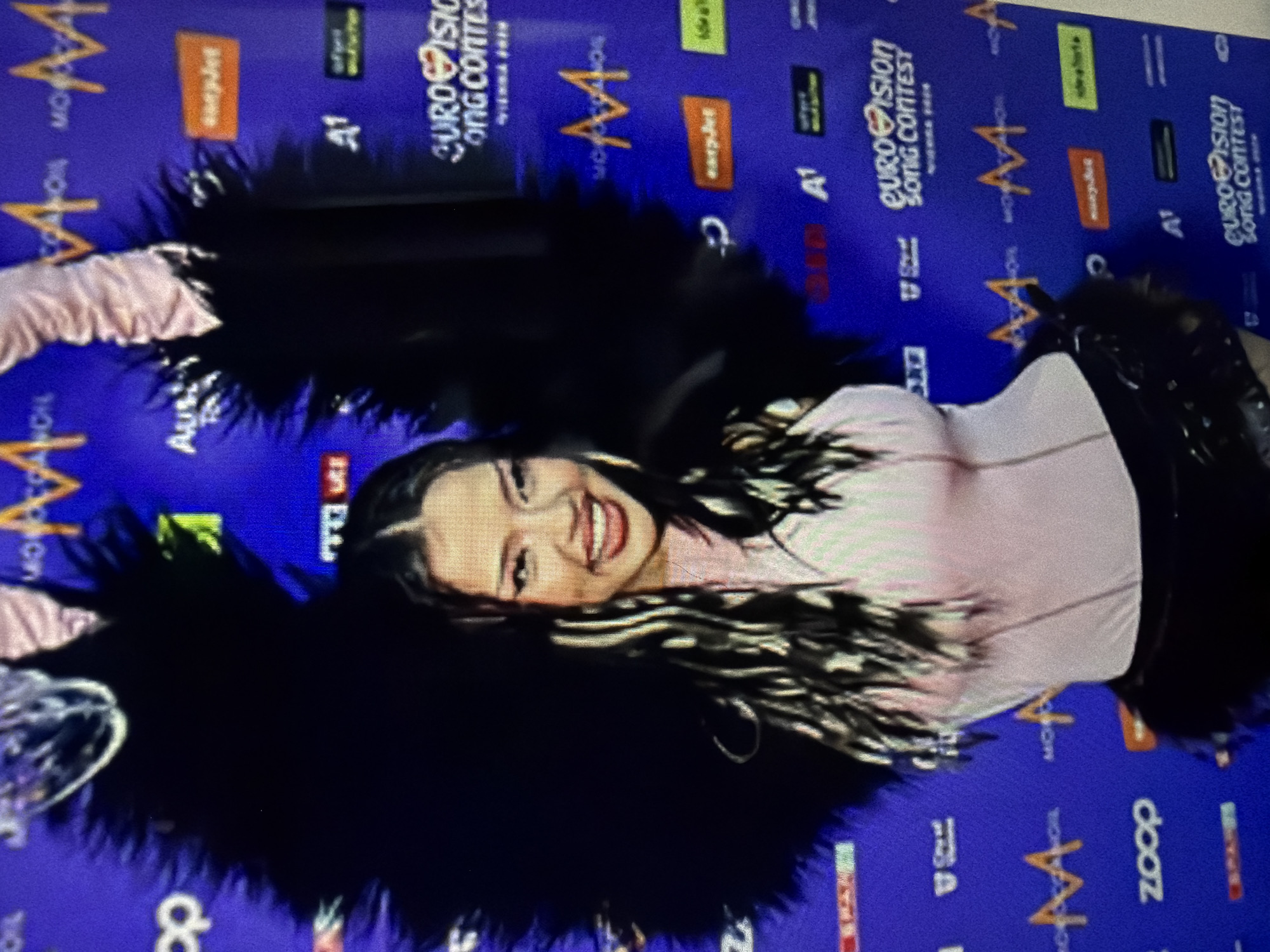 The image depicts a woman with dark hair and a light pink top posing on a blue carpet with various logos, including "A1" and "Eurovision Song Contest" printed on it. She is wearing a black, possibly leather, skirt and a large, black feather-like accessory around her shoulders. The background consists of various logos, indicating a promotional or event setting. There is no information to indicate the location is in Ruse, Bulgaria. The scene appears to be a red carpet or promotional event.
