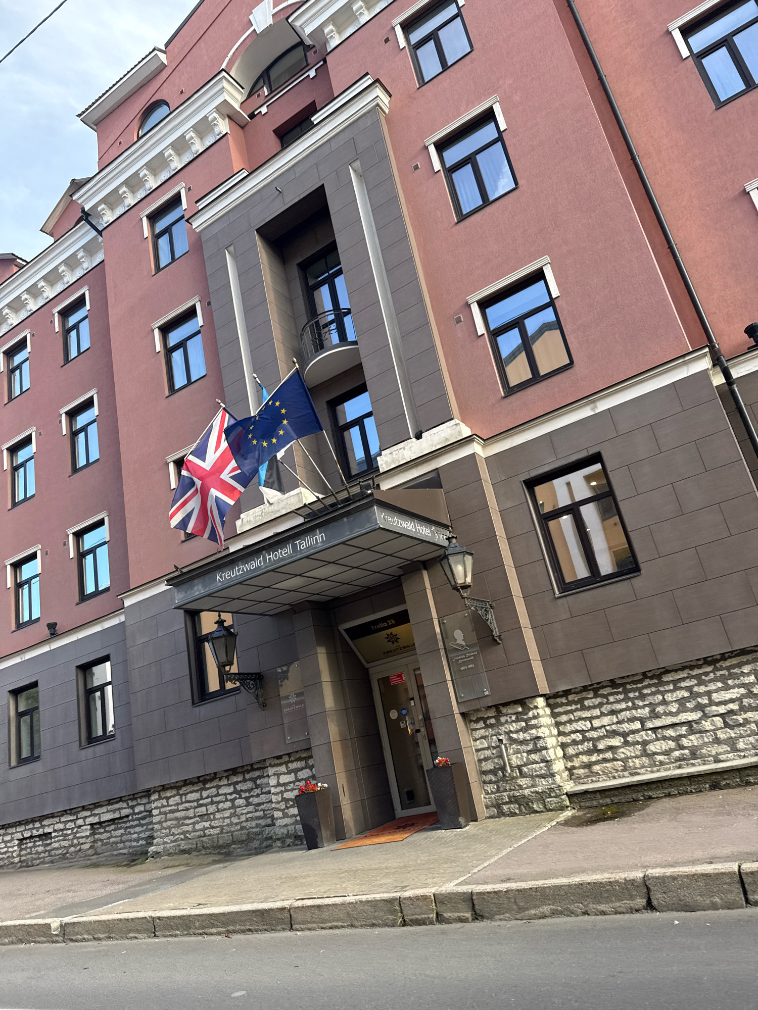 The image shows the facade of the Kreutzwald Hotel in Tallinn, Estonia. The hotel is a multi-story building with a pinkish-red exterior. It features numerous windows, some of which are reflecting the sky and surrounding buildings. Two flags are prominently displayed above the entrance: the Union Jack of the United Kingdom and the flag of the European Union. Below the flags, a sign reads "Kreutzwald Hotell Tallinn". The entrance to the hotel is visible, with a glass door and a mat on the ground. Adjacent to the entrance, there is a stone-covered lower level of the building. The scene is set on a street with a sidewalk and a curb. The overall impression is that of a well-maintained hotel in an urban setting.