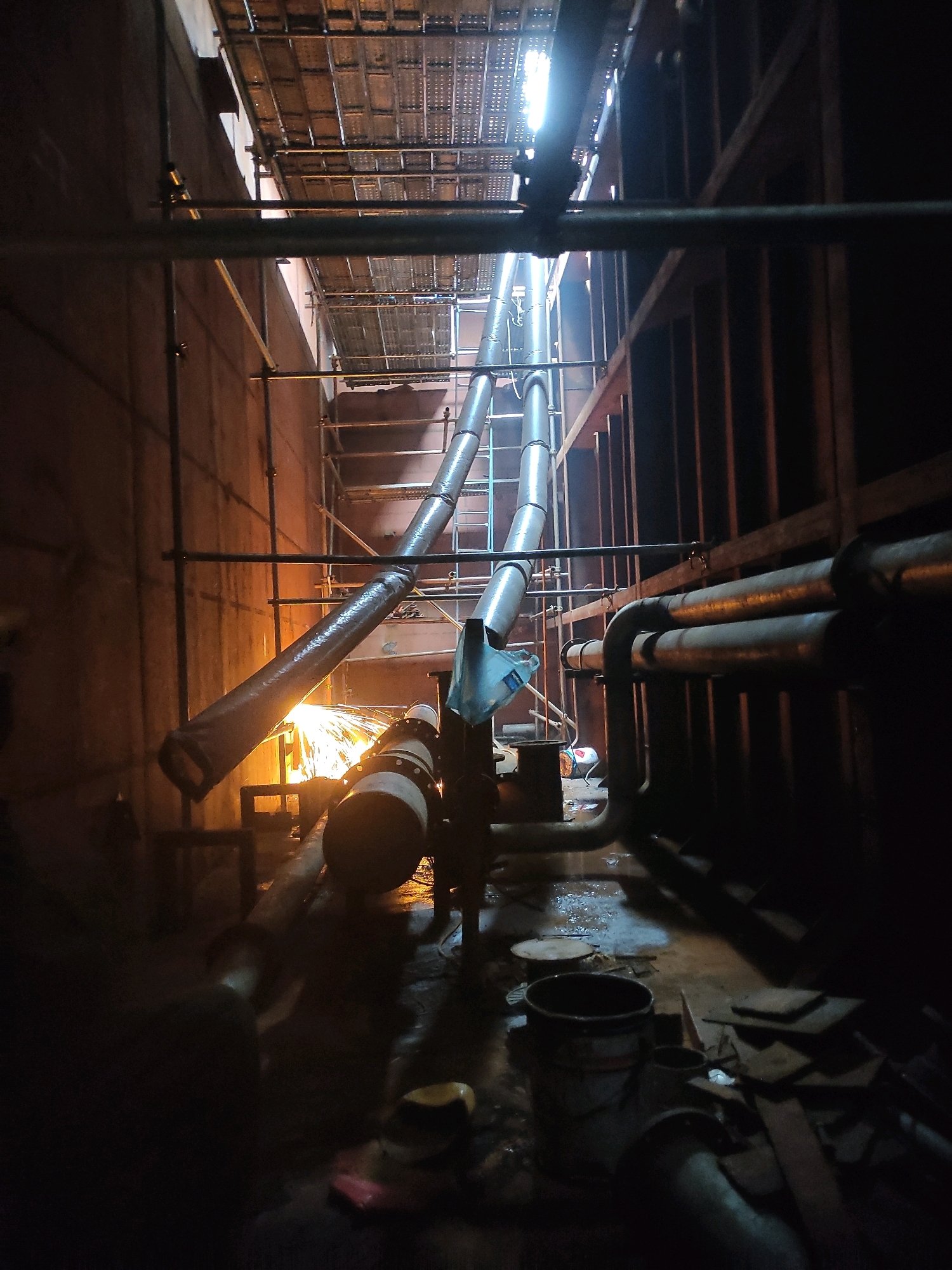The image depicts an industrial setting, likely within a construction or maintenance area in Batam, Indonesia. The scene is dominated by large, grey pipes that run diagonally across the frame, some appearing to be insulated. Sparks are visible emanating from the lower section of one of these pipes, suggesting welding or grinding activity is in progress. Scaffolding is erected along the walls of the space, which are dark and appear to be constructed of concrete or metal. Various other pipes, some rusty, are also present on the right side of the image, running horizontally. The floor is wet and appears to be littered with debris, including buckets and pieces of wood or metal. A bright light source is visible at the top of the image, illuminating the upper sections of the pipes and scaffolding. The overall impression is of an active, possibly hazardous, industrial work environment.