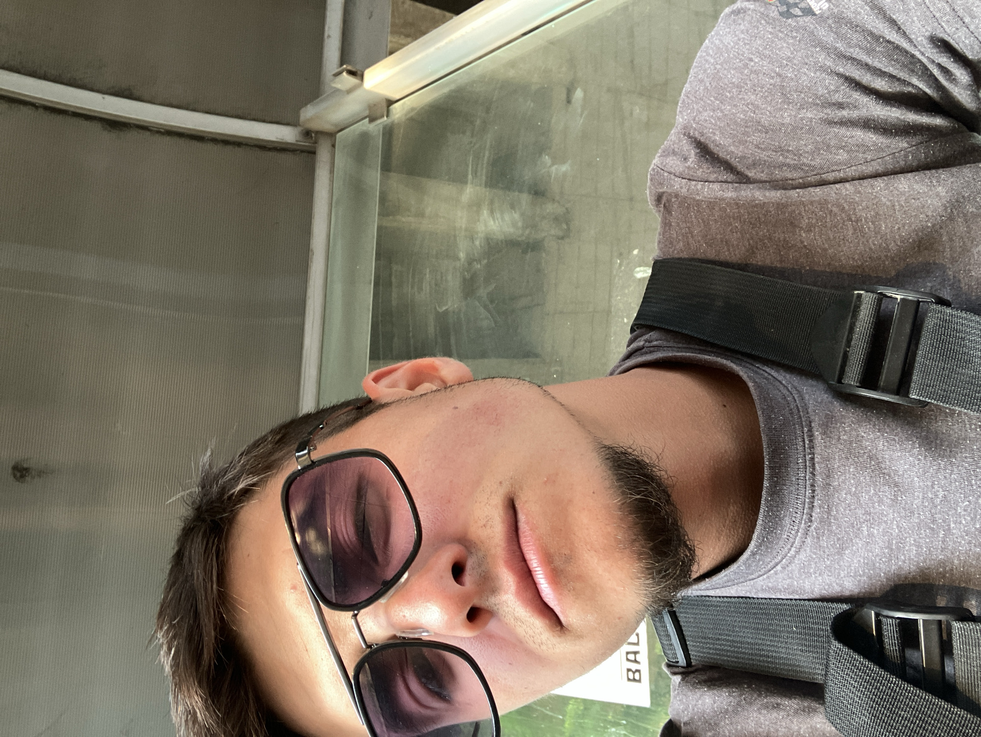 A man wearing sunglasses and a harness is inside a glass structure. He has dark hair and a beard. He is wearing a gray t-shirt. A sign with "BAL" is partially visible. The location is in Plovdiv, Bulgaria.