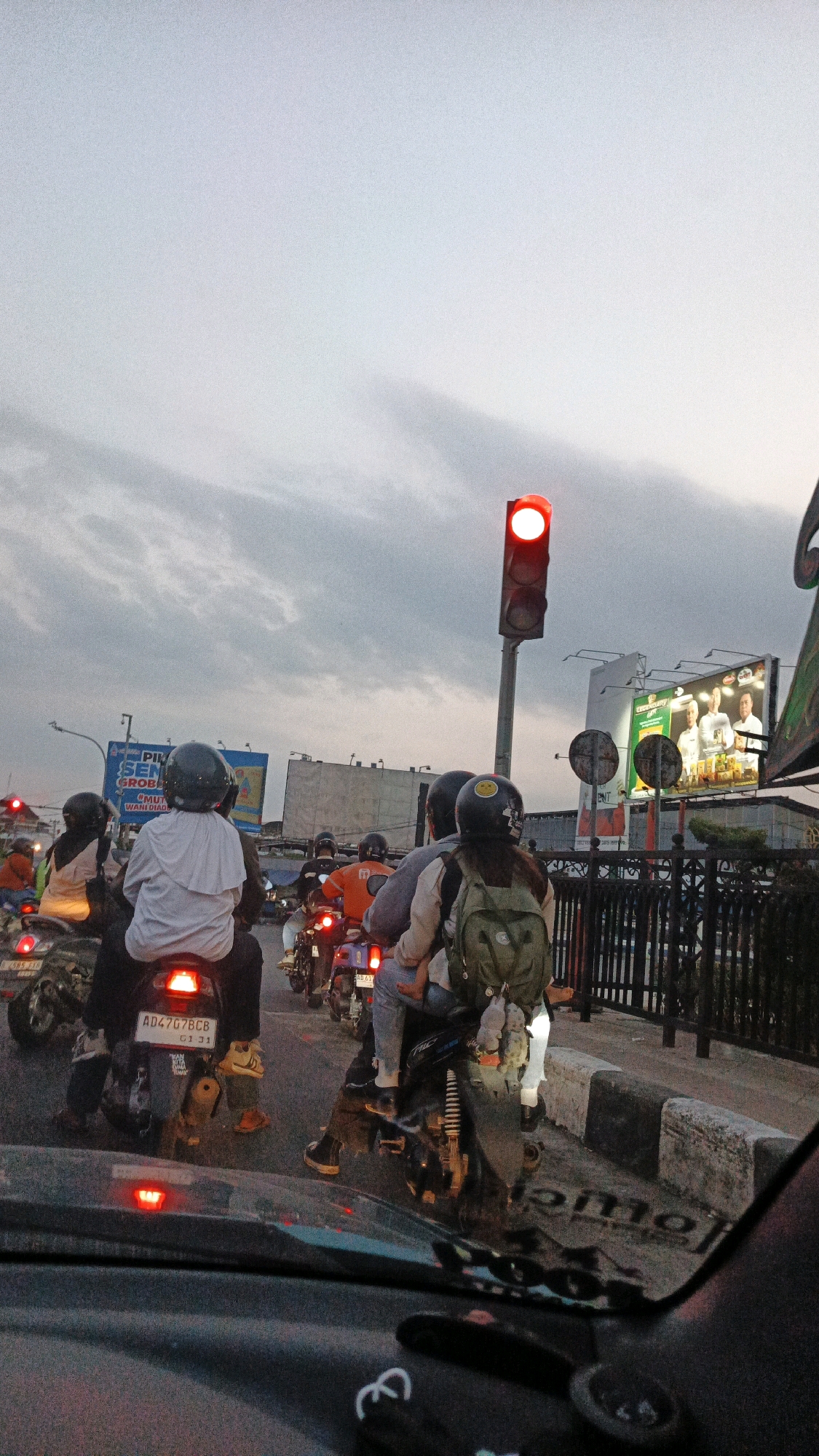 In Surakarta, Indonesia, on a street filled with moderate traffic, a group of motorcyclists are stopped at a red traffic light. The sky is overcast with dramatic clouds, suggesting either dusk or an approaching storm. The foreground is dominated by the windshield of a car, reflecting the road and traffic lights. The motorcyclists, wearing helmets for safety, are diverse in age and gender. One motorcyclist in the middle wears a white shirt and a black helmet, while another behind them has a black helmet with a smiley face sticker. A female motorcyclist in the right foreground wears a green backpack and a black helmet. The traffic light is illuminated red, indicating a stop. In the background, billboards advertising various products are visible, and a black metal fence lines the side of the road. The overall scene captures a typical urban traffic moment in Surakarta.
