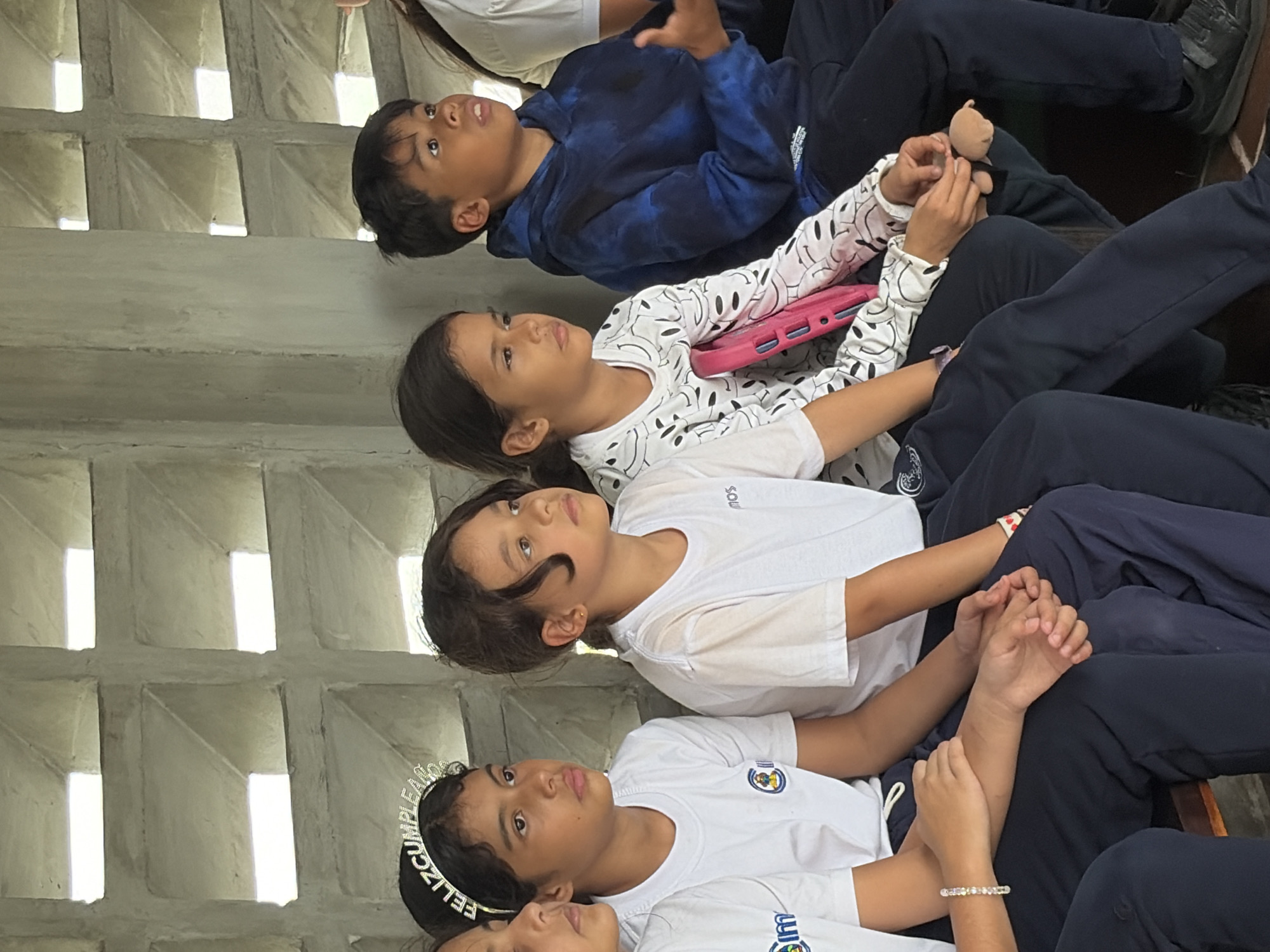 The image shows four children sitting in a row, possibly on a bench or steps. They are looking upwards and to the right. The first child, a boy, is wearing a blue patterned hoodie. The second child, a girl, is holding a pink tablet and wearing a white long-sleeved shirt with black patterns. The third child, a girl, is wearing a plain white t-shirt. The fourth child, also a girl, is wearing a white t-shirt and a headband with the word "FELIZ CUMPLEAÑOS". All the children are wearing dark pants or leggings. The background shows a textured, light-colored wall with a pattern of square openings, letting in light. The location is suggested to be Ciudad Bolivar, Venezuela. The children are sitting in close proximity, with their legs and hands overlapping or touching, suggesting they may be interacting or engaged in a shared activity.