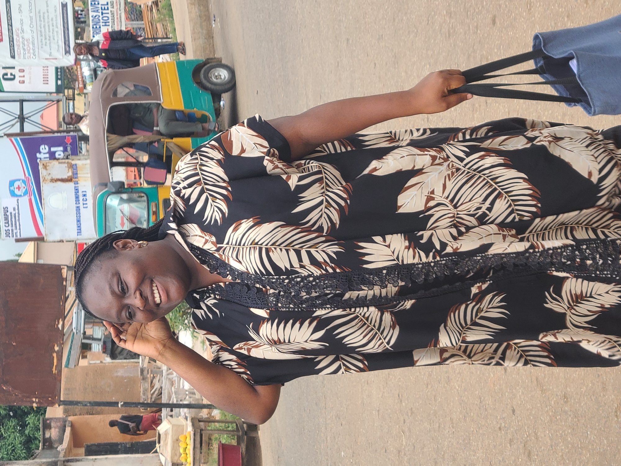 The image shows a woman standing on a city street, smiling at the camera. She is wearing a black dress with a beige and brown leaf print and holding a blue tote bag. Behind her is a "keke", a yellow and green taxi-tricycle, with a few people in it. In the background are buildings and street signs, one of which reads "FRIENDS AVENUE HOTEL". Another sign shows the acronym TCN, for Transmission Company of Nigeria.
