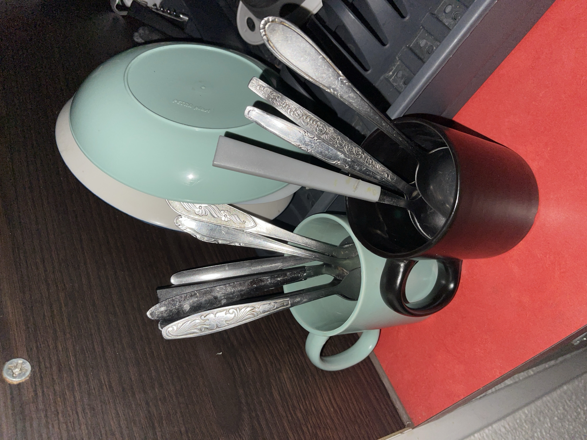 The image displays a close-up view of kitchen items stored in a cabinet. In the foreground, two mugs are visible: a light teal mug to the left, filled with several spoons and what appears to be a knife, and a black mug to the right, also containing spoons. Behind the mugs, a stack of plates, one light teal and one white, are propped up. To the right of the plates, a gray dish rack is partially visible, holding more utensils. The background consists of a dark brown wooden surface, possibly the inside of a cabinet, with a single silver screw visible on the upper left. The overall scene is indoors, on a red surface that could be a countertop or shelf liner. The image does not contain any contextual information indicating a location in Pilsen, Czechia.