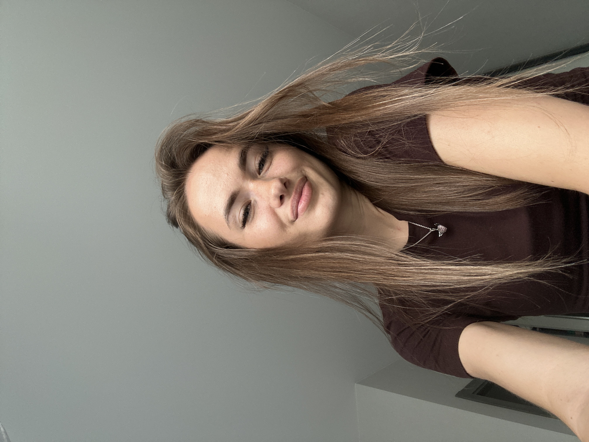 A young woman is captured from a low angle, looking up towards the camera. She has long, light brown hair that is slightly messy and falls around her shoulders. Her eyes are dark and her eyebrows are well-defined. She has a slight smile on her face, with her lips slightly parted. She is wearing a dark brown t-shirt and a silver necklace with a small pendant. The background is a plain, light grey wall, with a hint of a white corner in the upper left. The lighting appears to be coming from above and slightly in front of her. The overall impression is a casual, candid selfie.