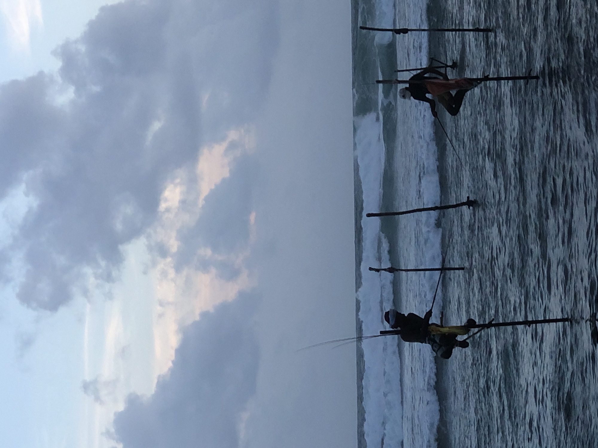 The image depicts two stilt fishermen in the shallow waters off the coast of Kathaluwa, Sri Lanka, during overcast daylight. The fishermen are perched on tall wooden stilts, each holding a fishing rod. The ocean is rough with visible waves crashing. The sky is filled with gray and white clouds, with hints of pale pink and blue. The overall scene is somber and highlights the traditional fishing practice unique to the region.
