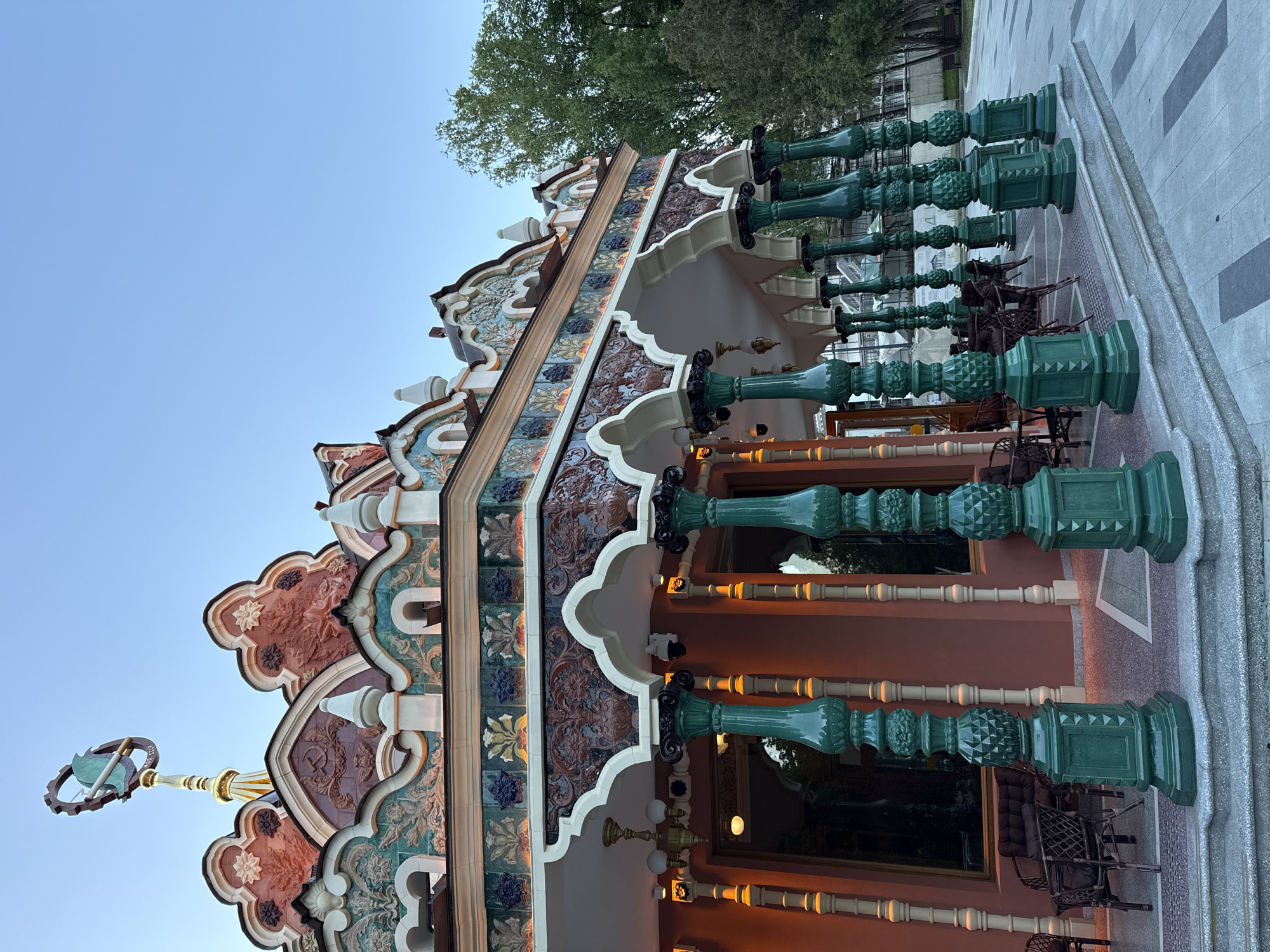 This image captures the richly decorated facade of a building, likely a historic or culturally significant structure, against a clear blue sky. The building exhibits an ornate architectural style, possibly Art Nouveau or Moorish revival, with a distinctive roofline featuring numerous curvilinear elements, finials, and intricate tile work.
The facade is adorned with vibrant, multi-colored ceramic tiles depicting floral motifs, geometric patterns, and embossed designs in shades of teal, brown, red, and cream. The base of the building appears to be a reddish-brown. Below a series of arches, several tall, dark green, highly decorative pillars or lamp posts with various textured and patterned sections (fluted, diamond-cut, smooth) line the front, suggesting an entrance or outdoor seating area. Behind these pillars, chairs and tables are visible, indicating a potential outdoor cafe or restaurant.
On the far left, atop a golden spire, a prominent decorative emblem features a hammer and sickle, a symbol associated with the Soviet Union, encircled within what appears to be a gear or wreath. The letters "СССР" (USSR) are clearly visible around this emblem. The sky is a light, clear blue, suggesting a daytime shot, possibly late afternoon given the soft lighting and the warm glow from some internal or external building lights that are beginning to illuminate. Trees are visible in the background, further indicating an outdoor setting. The ground features patterned paving leading up to the building. The overall scene exudes a sense of historical grandeur and intricate craftsmanship, characteristic of Moscow, Russia.