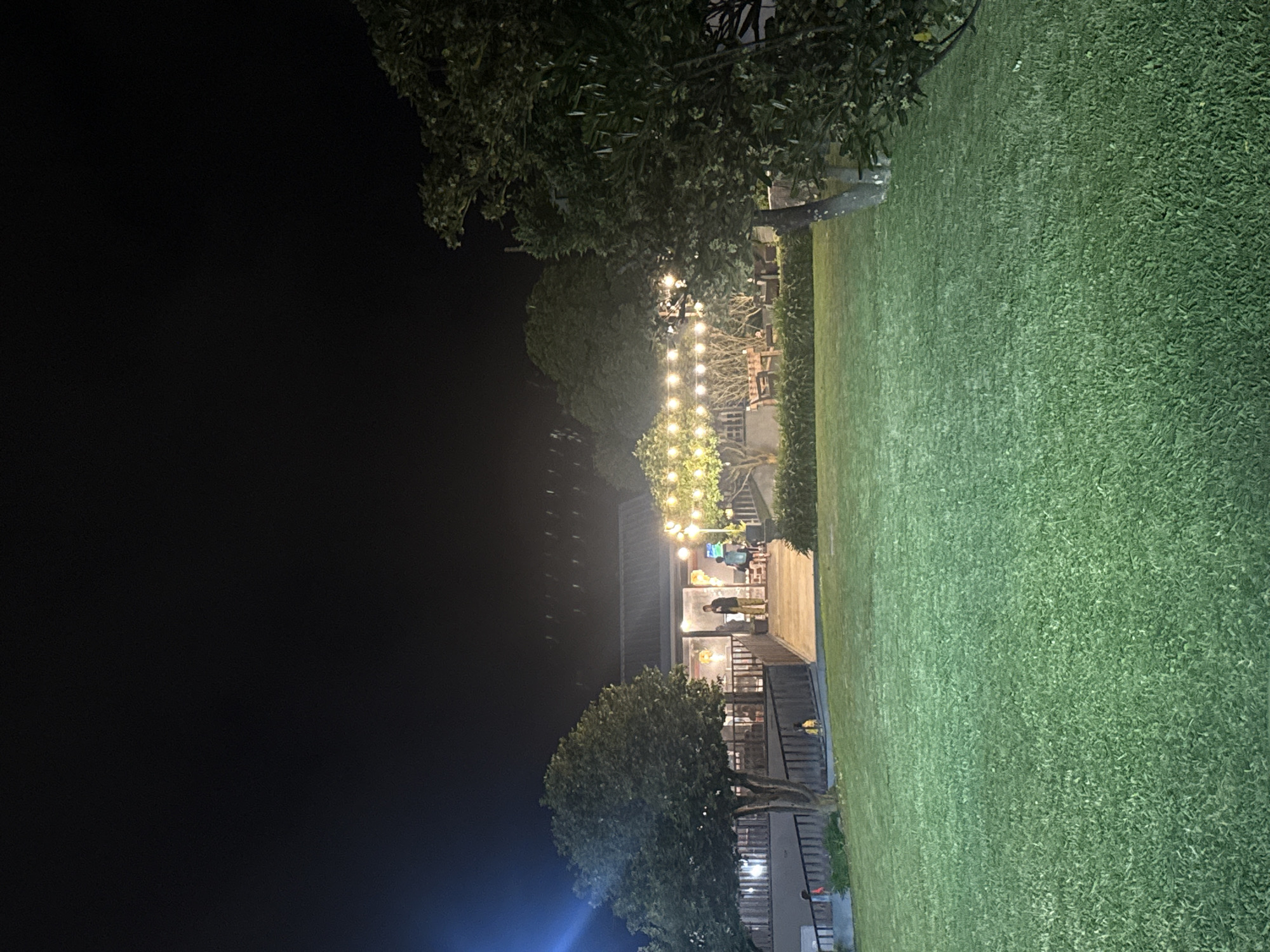 The image depicts an outdoor scene at night in Moreira, Portugal. A well-manicured lawn occupies a significant portion of the right side. A structure with multiple levels is visible, illuminated from within and by strings of decorative lights. People are present on the lighted patio of the building. Trees are interspersed throughout the scene, framing the structure and adding to the outdoor ambiance. The left side of the image is dark, contrasting with the illuminated building and lawn.