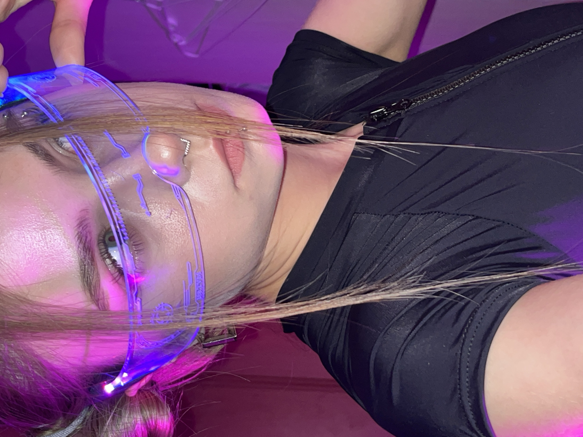 A close-up shot depicts a fair-skinned individual with light brown hair. They are wearing futuristic, clear glasses with blue neon lighting on the frames. The person's left eye is visible, reflecting the blue light. They have a lip ring on their upper lip. The person is wearing a black, form-fitting garment with a visible zipper on the neckline. The background includes shades of pink and purple, suggesting neon lighting effects. There is no indication of the location being the city of Фоки, Russia.