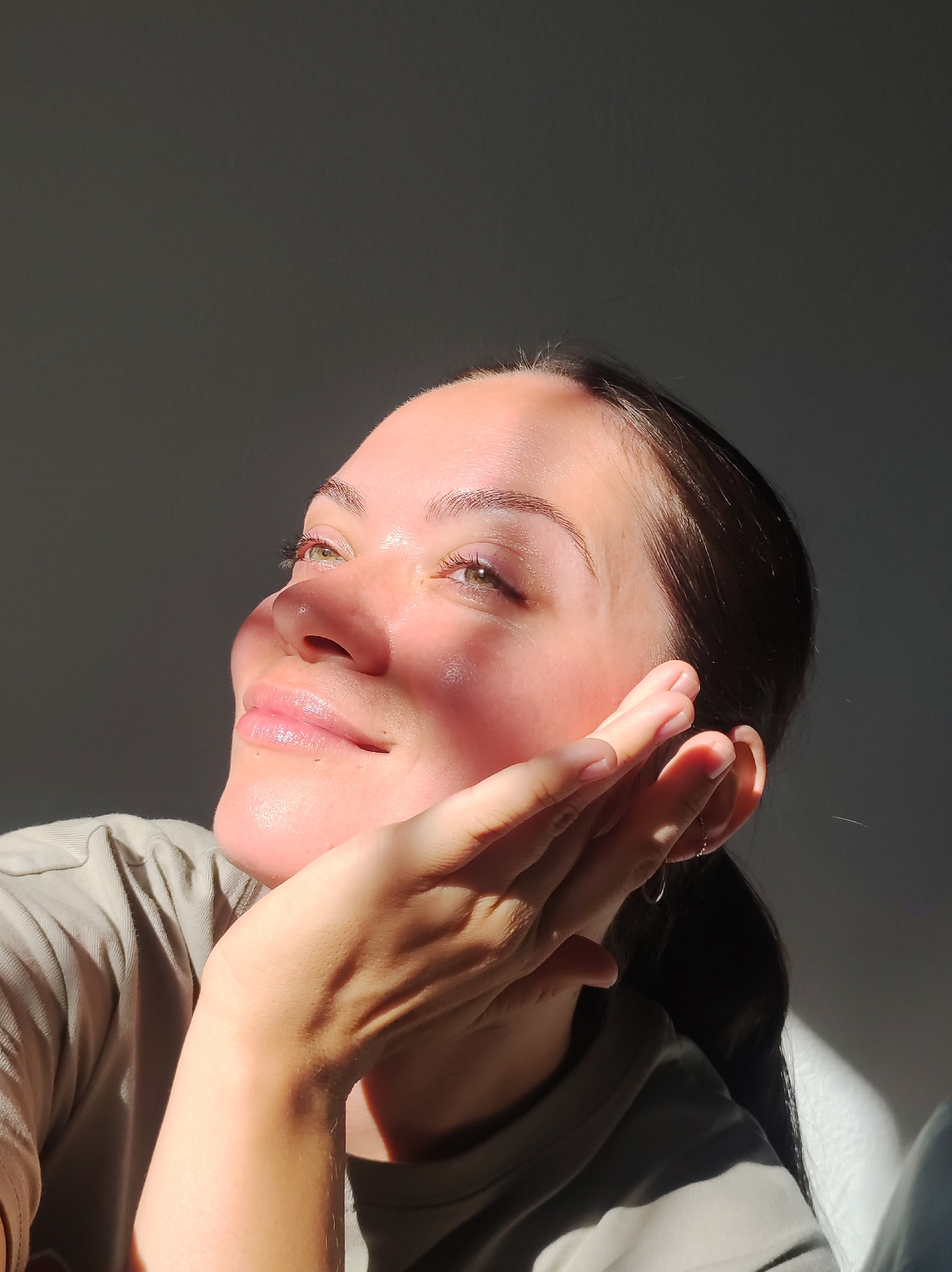 A close-up, medium shot captures the face of a young woman, bathed in warm, golden sunlight. The light casts dramatic shadows across her face, highlighting her features and giving the image a serene and contemplative mood. Her eyes are closed, and a gentle smile plays on her lips, suggesting a sense of peace or contentment. She rests her chin on her hand, her fingers delicately touching her cheek. Her hair is pulled back, revealing her ear, adorned with a small hoop earring. She wears a light-colored, textured t-shirt. The background is a plain, dark gray wall, which serves to accentuate the subject and the play of light and shadow on her skin. The strong directional sunlight indicates it is likely either early morning or late afternoon.