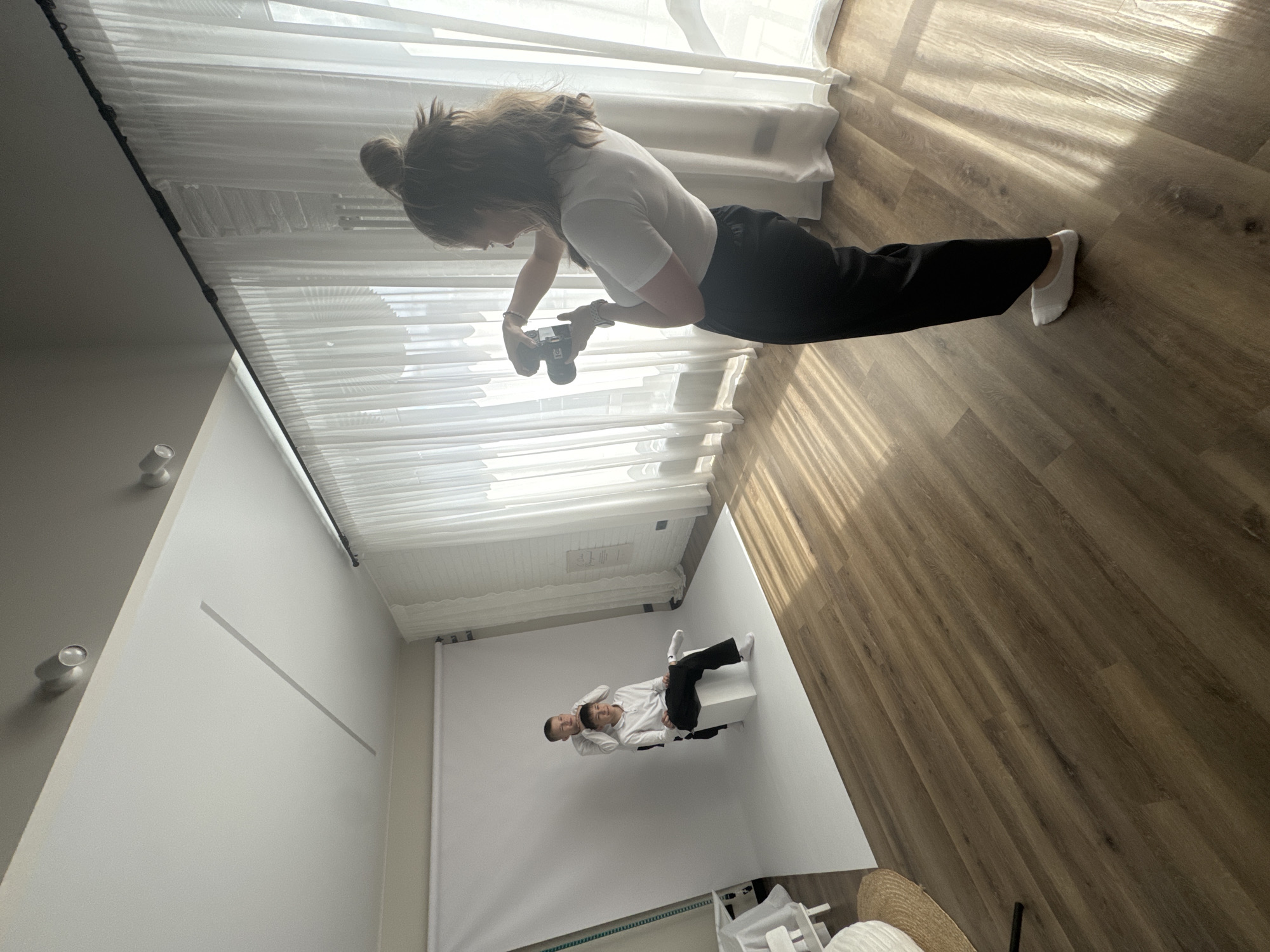The image shows an indoor scene in Izhevsk, Russia. A woman with her hair in a bun is taking a photo with a digital camera. She is wearing a white t-shirt, black pants, and white socks. She is standing on a light brown wood floor near a window with white curtains.
In the background, a white backdrop is set up. Two boys are sitting on a white cube in front of the backdrop. One boy is leaning back and smiling, and the other boy is behind him. They are both wearing white shirts and black pants.
Near the bottom of the image, there is a white storage rack and a straw hat. A grey line is visible on the wall in the background. The lighting is bright and natural.