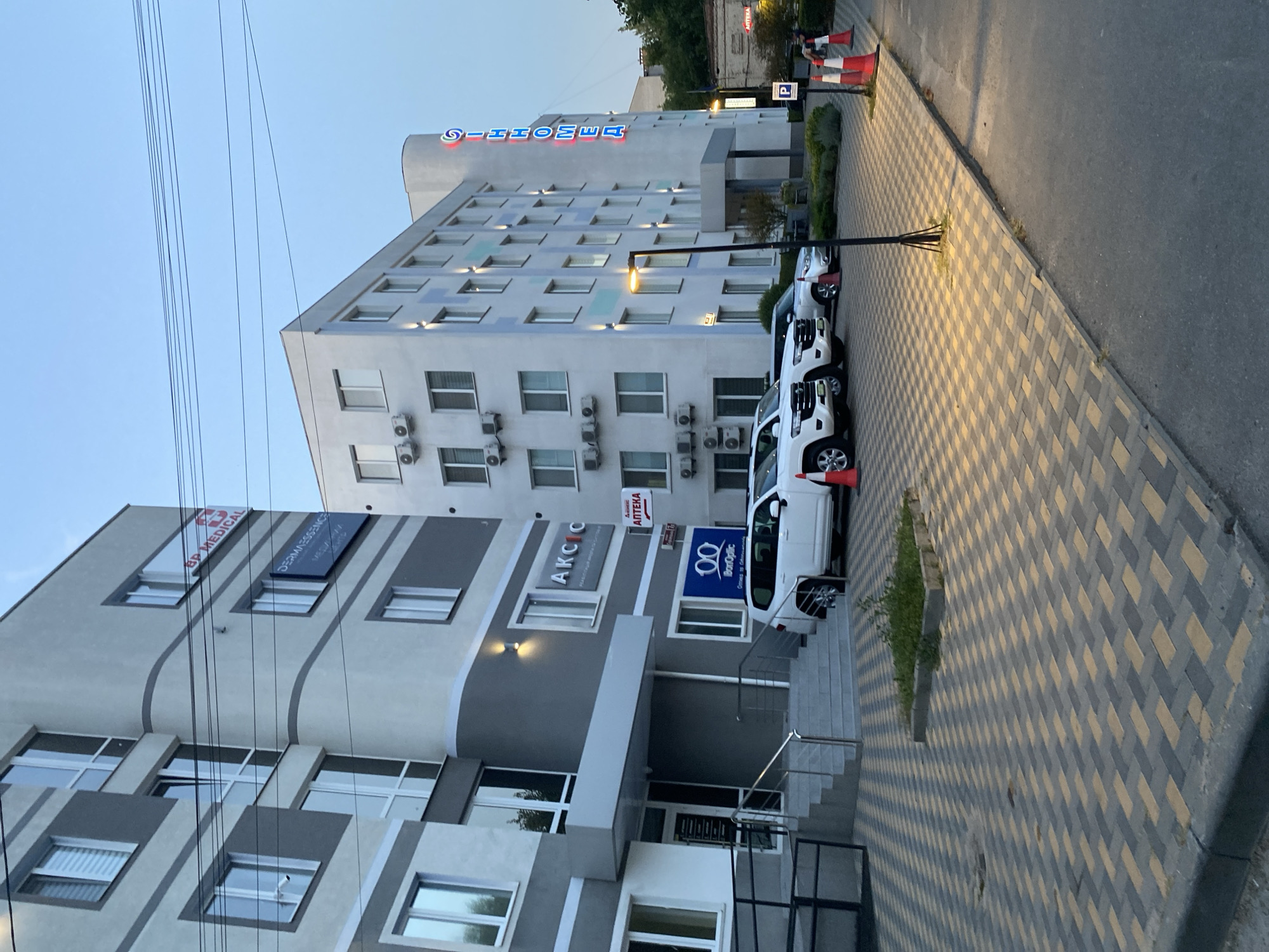 The image shows a street view in Vinnytsia, Ukraine, featuring a modern medical complex. Two multi-story buildings, predominantly white and grey, dominate the scene. One building, on the left, has a curved section and displays signage for "BP MEDICAL" and "DERMAESSENCE." The other building, on the right, features a prominent neon sign reading "SINHOMED" in blue and red letters. The street is paved with a distinctive pattern of grey and yellow interlocking bricks. Several white SUVs are parked in front of the buildings, and traffic cones are visible on the right side of the image. The sky is a clear, pale blue, suggesting it is either early morning or late evening. The overall impression is of a clean, well-maintained urban environment focused on healthcare services.