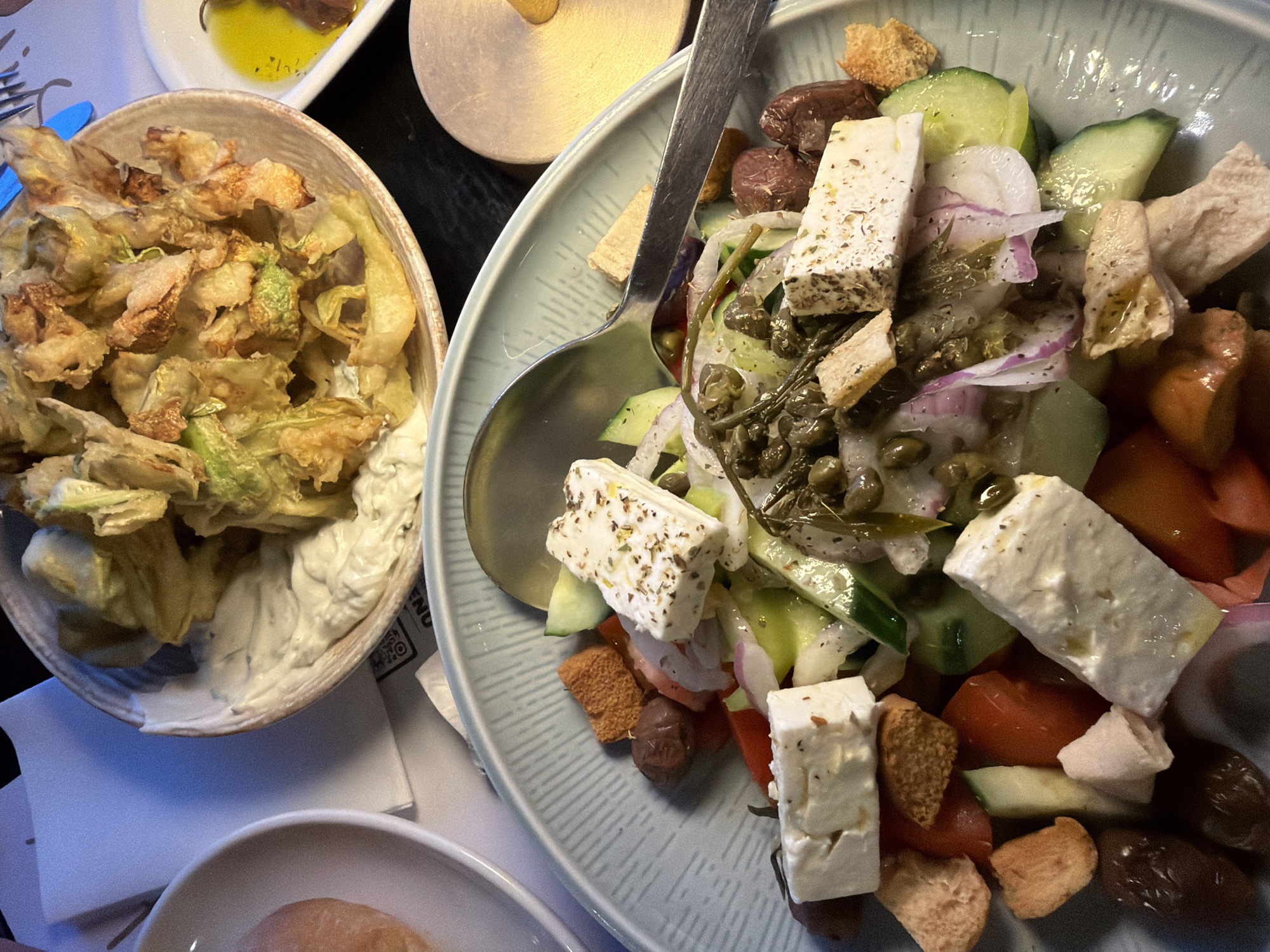 The image captures a vibrant and appealing dining scene, likely in a restaurant setting in Makri, Greece. Two main dishes are prominently featured on a dark table surface, illuminated by warm artificial lighting, suggesting an evening meal.
On the right, a large light blue-green bowl, characterized by a subtle textured pattern, holds a hearty Greek salad. The salad is composed of generous rectangular cubes of white feta cheese, visibly seasoned with dried herbs. Other ingredients include thick slices of green cucumber, thinly sliced red onions, red tomato chunks, dark Kalamata olives, and golden-brown croutons. Small dark green capers or caper berries are also scattered throughout the salad, which appears to be dressed with oil and herbs. A silver spoon is resting in the salad.
To the left, a rustic, light-brown ceramic bowl contains a pile of golden-brown fried vegetables. These appear to be fried zucchini or possibly zucchini blossoms, exhibiting a mix of crispy textures and some greener parts. The vegetables are served atop a creamy white dipping sauce, likely tzatziki or a similar condiment.
In the background, a small white rectangular dish containing a pool of yellow olive oil is partially visible. At the bottom of the frame, part of another white bowl with a light-brown bread roll or bun can be seen. A white paper or napkin is visible beneath the fried vegetable bowl, displaying a small, illegible QR code. The overall scene conveys a delicious and traditional Greek meal being enjoyed.