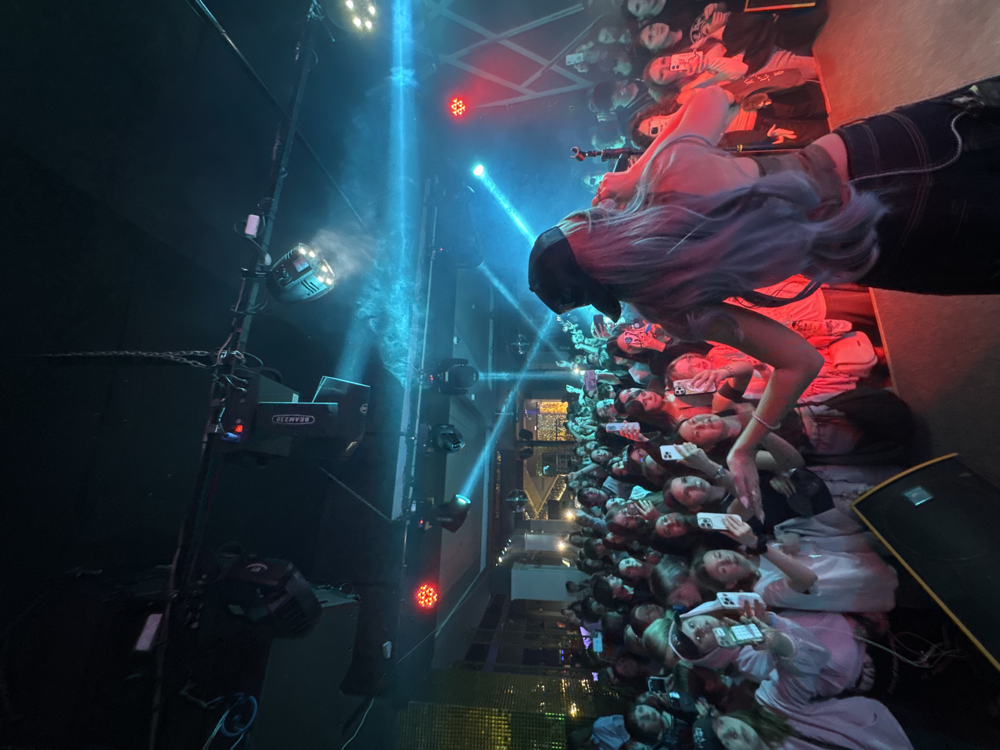 A crowd of people is gathered at a live event, likely a concert, in Yuzhno-Sakhalinsk, Russia. Most individuals are holding up their mobile phones, apparently recording or photographing the performer. The stage is illuminated with bright blue spotlights and red stage lights. A person with long, light blue hair is on stage. Stage lighting equipment and other objects are visible. The area is illuminated, but some portions are obscured by shadow or bright light. A large speaker is positioned at the bottom right.