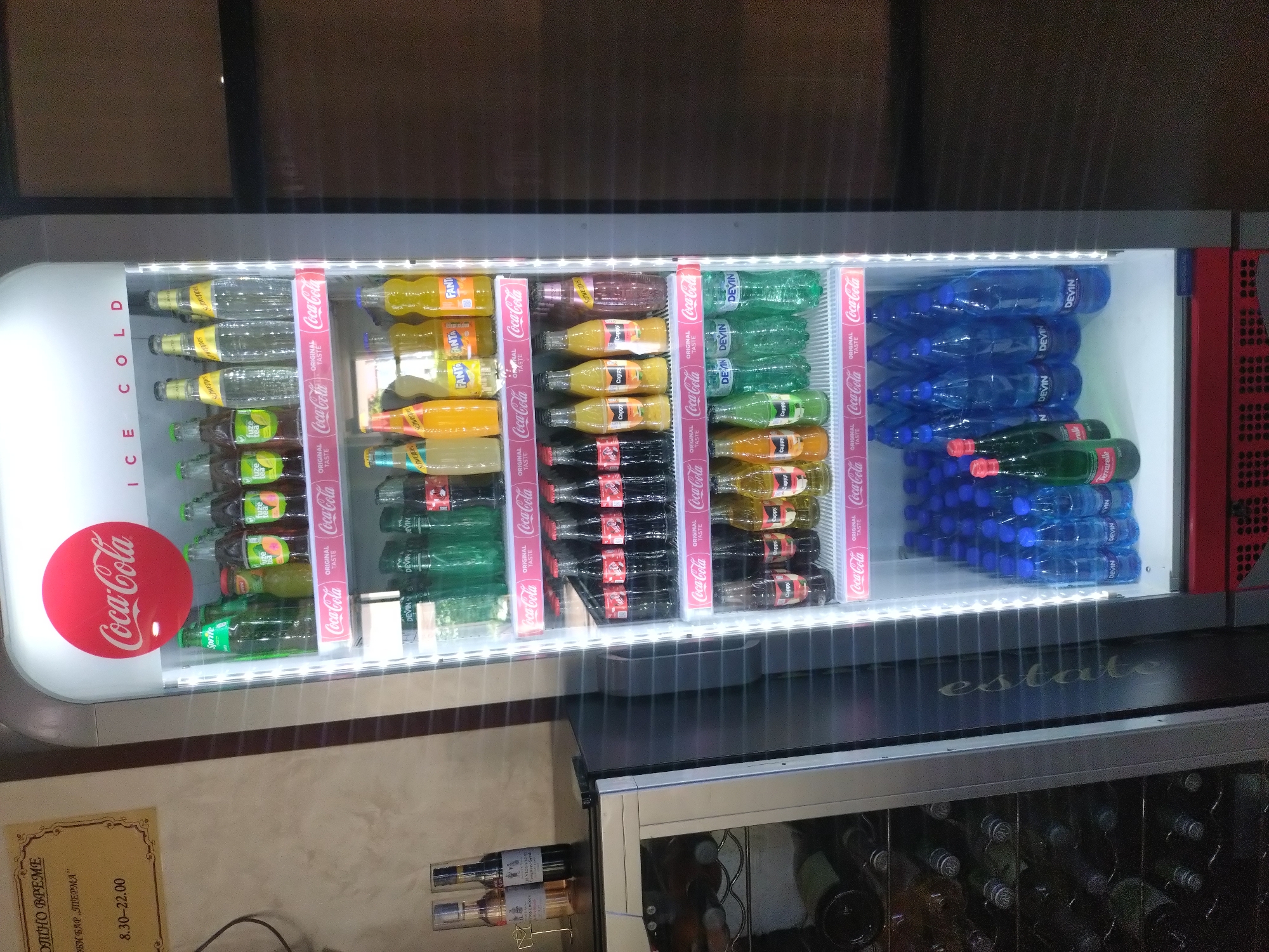 The image shows a well-stocked beverage refrigerator, likely in a bar or shop, as indicated by the "OPEN TIME" and "BAR" text in Bulgarian on a sign in the lower left. The refrigerator is brightly lit from within, showcasing a variety of bottled drinks. The primary subjects are the numerous bottles of soft drinks and water, neatly arranged on shelves. Brands like Coca-Cola, Sprite, Fanta, Schweppes, Fuze Tea, and Devin water are clearly visible. The "ICE COLD" text on the side of the refrigerator emphasizes its function.
In the background, a dark, indistinct wall and ceiling are visible, suggesting an indoor setting. The lower part of the image shows another cabinet, possibly a wine cooler, with bottles inside. The overall scene is one of commercial display and abundance of refreshments. There are no people visible in the image. The lighting suggests it could be any time of day, as it's indoors. The text "estate" is subtly visible on a dark surface below the refrigerator. The sign in Bulgarian, "ОТНО ВРЕМЕ ОБИ БАР „ПЕРМА" 8.30-22.00", translates to "OPEN TIME BAR „PERMA" 8.30-22.00", indicating the operating hours of the establishment.