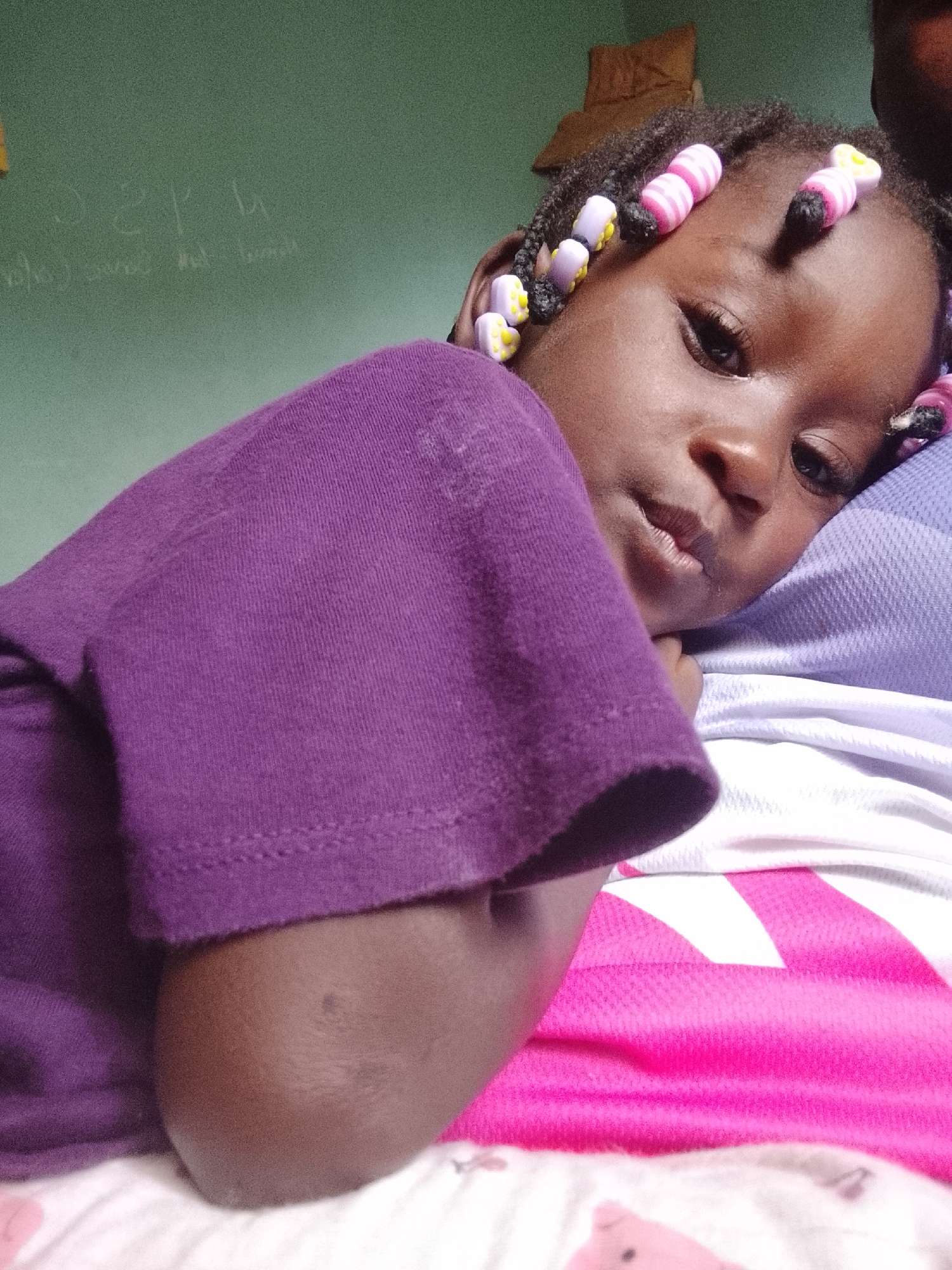 A young child with dark skin is captured in a close-up, lying down indoors, likely in a bedroom. The child, wearing a plain purple short-sleeved t-shirt, is positioned with their head resting on a light blue or purple patterned pillow. Their right arm is bent, with the elbow visible near the bottom right of the frame. Braids in the child's dark hair are decorated with colorful beads; some are pink and white striped, while others are yellow and white with a subtle pattern. The child's gaze is directed slightly downwards and to the side, conveying a calm or contemplative expression.
In the background, a light green wall is visible, featuring faint white markings that appear to be chalk or faded paint. While most of the writing is illegible, shapes resembling letters such as "S", "P", and "U" can be discerned, alongside other indistinct lines. Beneath the child, a vibrant pink and white patterned fabric, possibly a blanket or sheet with a geometric design, is visible, along with a white sheet displaying a very faint, light pink pattern, possibly of a cartoon animal's snout. The overall impression is a candid moment of rest or quiet observation.
