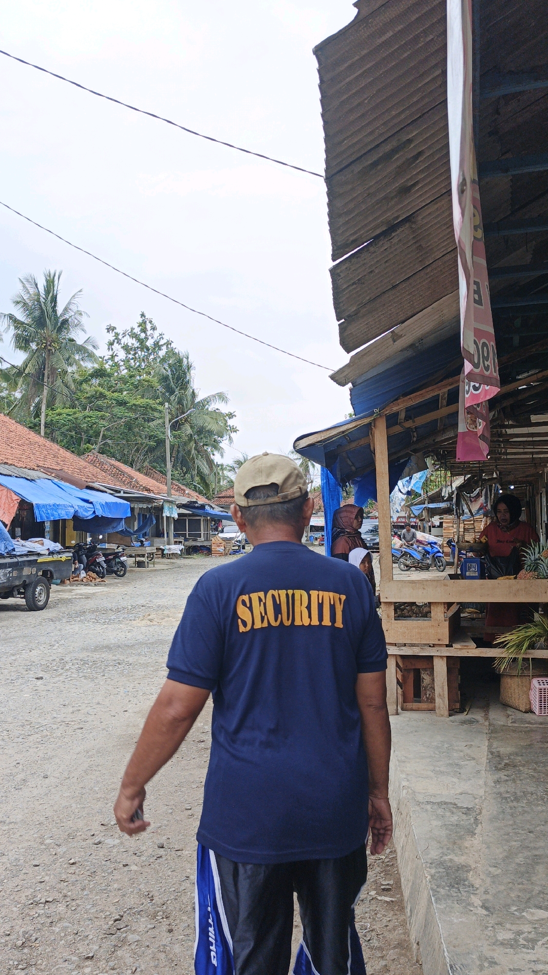 In Desa Karangbenda, Indonesia, a security guard wearing a navy blue t-shirt with the word "SECURITY" in yellow lettering is walking away from the camera on a gravel road. He is wearing a tan baseball cap and dark shorts with blue and white accents. In the background, there are several market stalls with corrugated tin roofs, some covered with blue tarps. Palm trees and lush green vegetation are visible behind the stalls, and a cloudy sky overhead suggests an overcast day. A pickup truck is parked to the left of the road. To the right, a woman is visible behind a market stall, appearing to be interacting with a customer.
