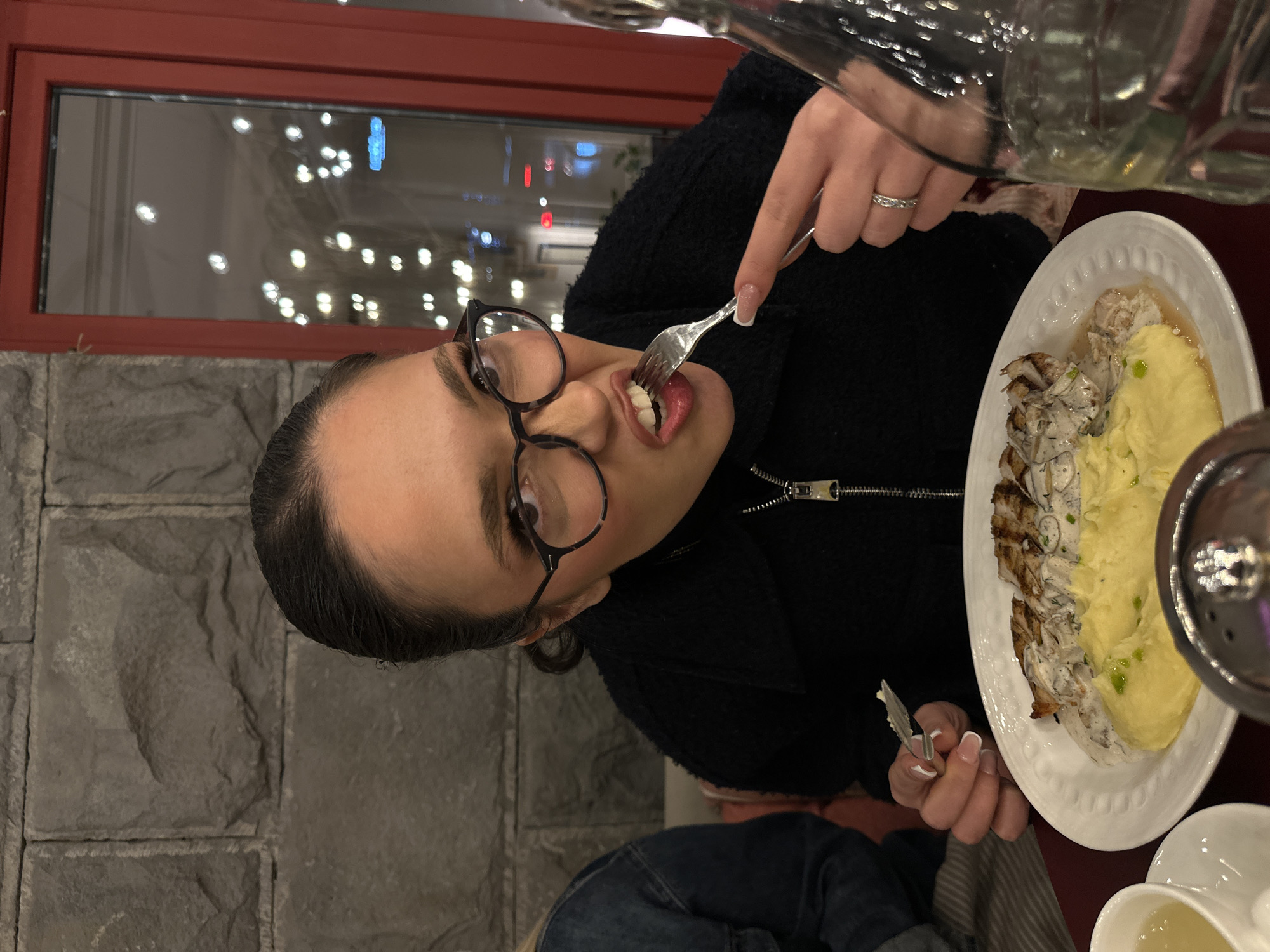 A woman with glasses is eating food at a restaurant. She is holding a fork with food in her mouth. The plate in front of her contains mashed potatoes and what appears to be sliced meat with a creamy sauce. The restaurant has a stone wall and a window in the background with blurry lights outside. There is a water pitcher in the upper right corner. This photo was taken in Yerevan, Armenia.