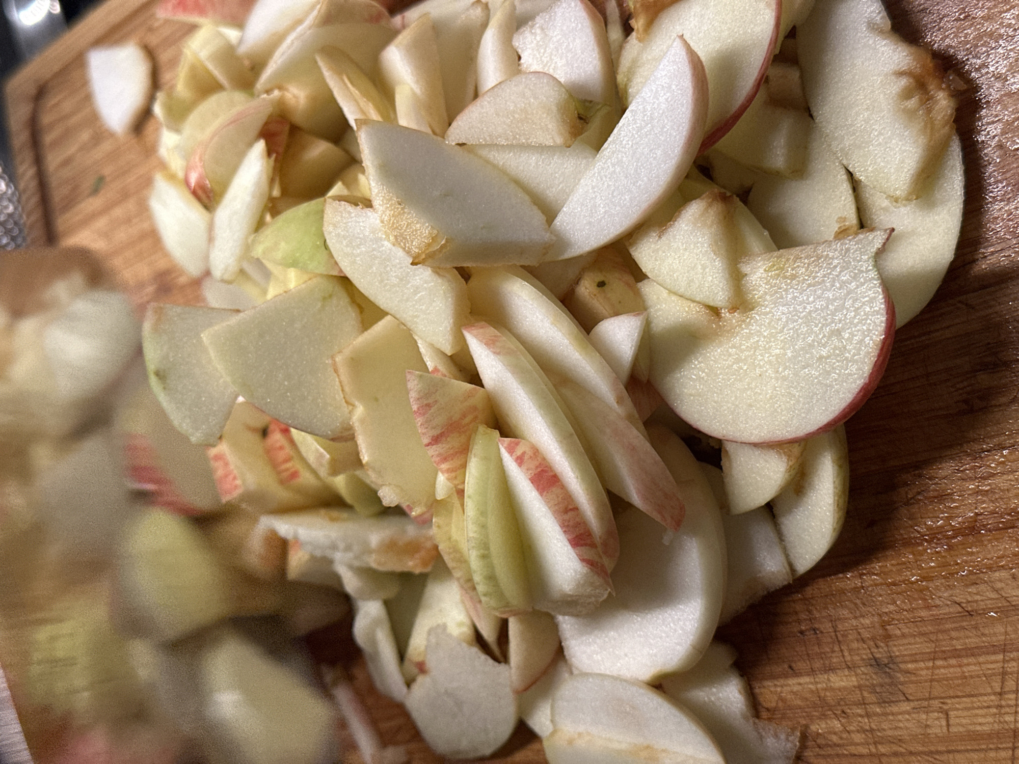 The image shows a pile of sliced apples on a wooden cutting board. The apples are cut into various shapes and sizes, with some pieces still showing the red skin. The cutting board has a visible wood grain texture and appears to be slightly wet in some areas. The lighting is warm, creating shadows and highlights on the apple slices. The background is blurred, but suggests an indoor kitchen setting. There are no people or animals visible in the image. The location of this scene in Nevinnomyssk, Russia cannot be determined from the visual information provided.