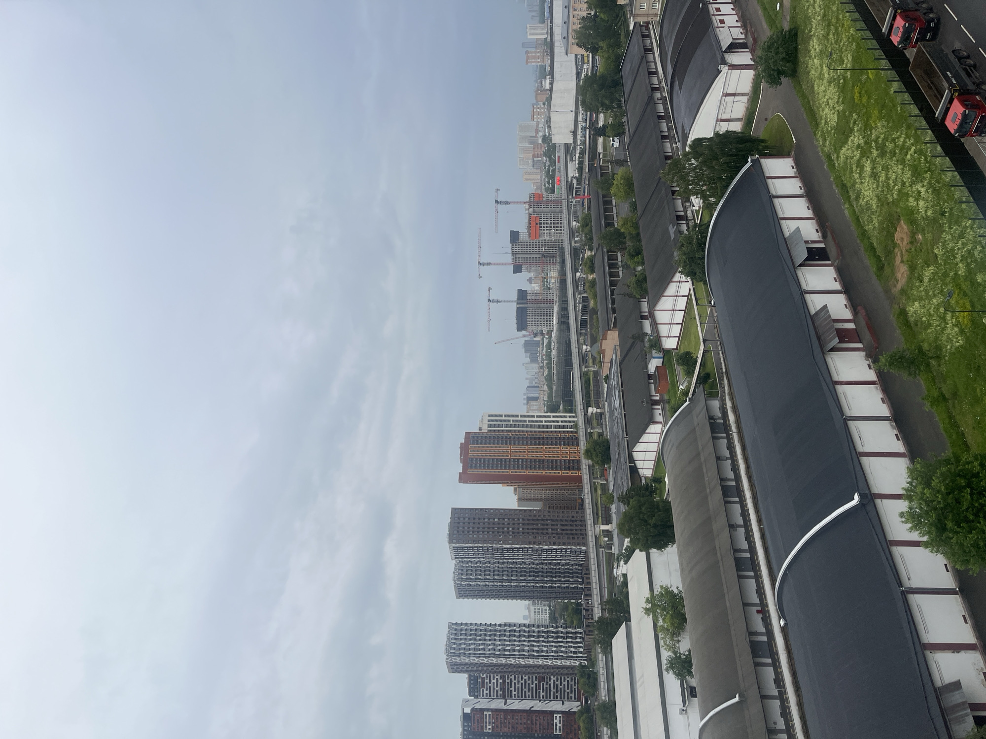 The image depicts an urban landscape in Mytishchi, Russia, characterized by a mix of residential high-rises, industrial buildings, and ongoing construction. In the background, a cluster of modern residential towers, varying in height and architectural style, are visible under a cloudy sky. Among them, a prominent red and orange colored building stands out. Further in the distance, construction cranes indicate active development of more high-rise structures. Closer to the foreground, a series of long, low-lying industrial or warehouse buildings with dark, arched roofs dominate the scene. These structures have white walls with red trim around doorways and windows. A elevated railway or highway passes between the residential and industrial areas. Greenery, including trees and grassy areas with scattered wildflowers, is present throughout the landscape. Two red dump trucks are parked on a road in the lower right corner of the image, suggesting ongoing logistics or construction activity. The overall atmosphere is overcast and slightly muted, typical of a cloudy day.