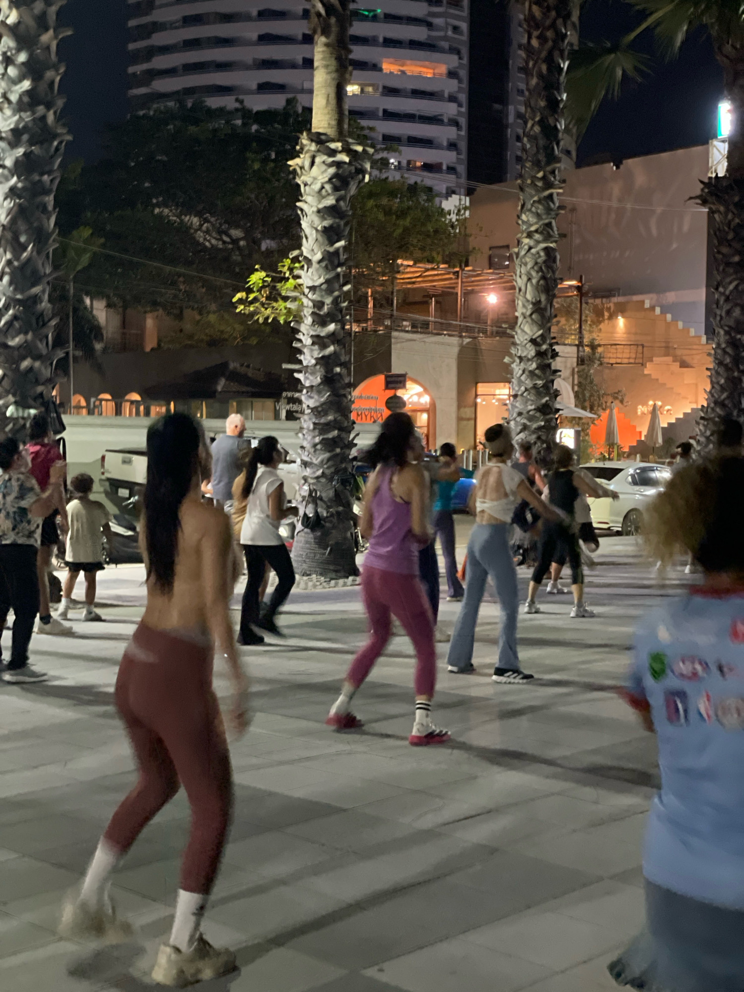 In Pattaya City, Thailand, a group of individuals is engaged in an outdoor exercise class at night. The scene is set against a backdrop of illuminated buildings and palm trees. Several participants, predominantly women, are captured in motion, wearing athletic attire such as leggings and tank tops. One individual in the foreground, with long dark hair, is wearing dark red leggings and white socks, her back to the camera as she performs a dynamic movement. To her right, another woman in a purple tank top and pink leggings is also in mid-stride. Other individuals, some in athletic wear and others in casual clothing, are dispersed throughout the frame, suggesting a lively fitness event. The ground is paved with square tiles, and shadows are cast by the palm trees and the artificial lighting. A white pickup truck is partially visible on the left side of the image, and a white car is parked further back. The overall atmosphere suggests a communal fitness activity taking place in a public urban space.