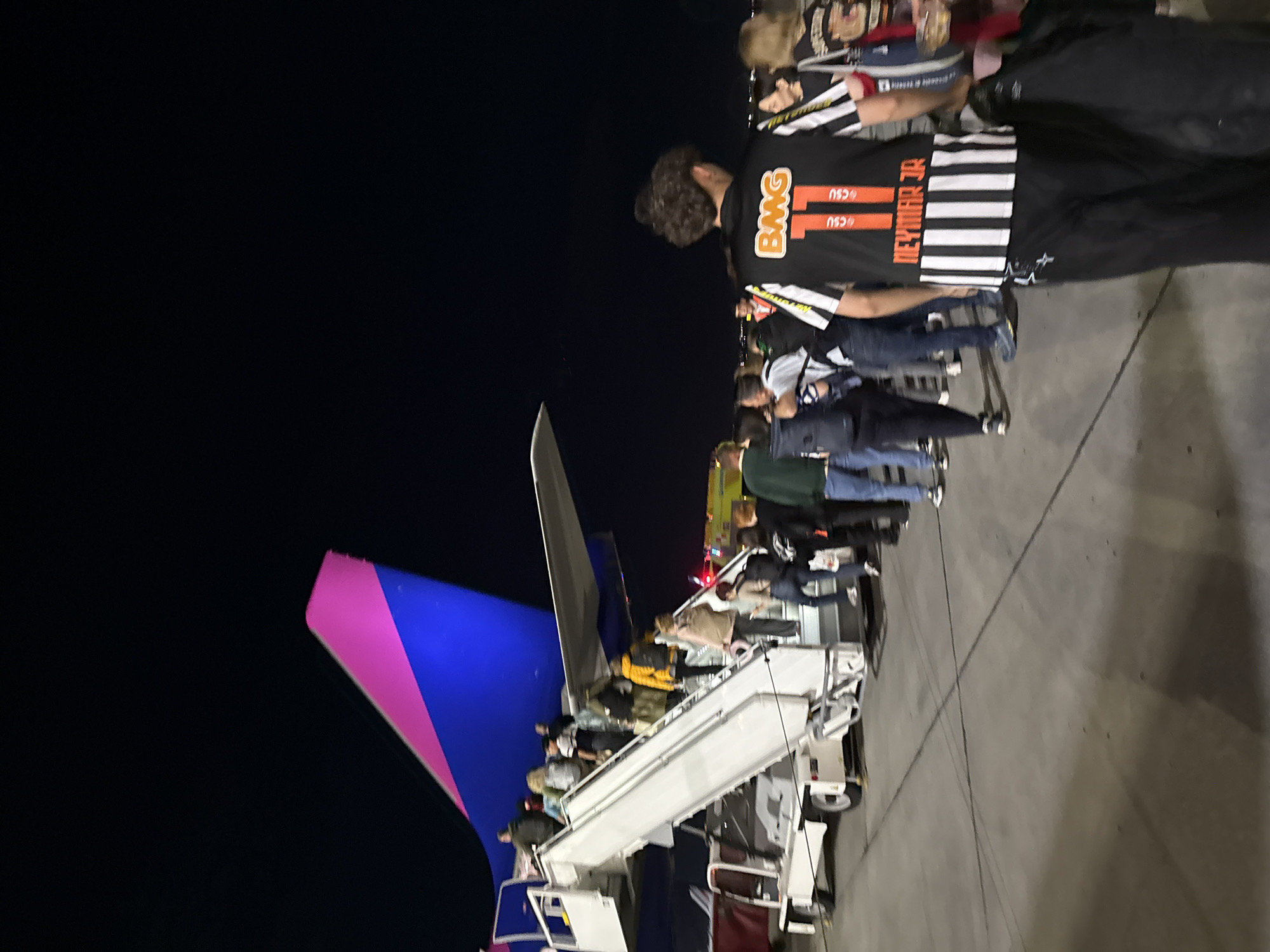 The image depicts a night scene at an airport tarmac in Yerevan, Armenia, showing people boarding an airplane. The sky is dark, indicating evening or night, with the scene illuminated by artificial ground lights.
In the foreground, a person is seen from behind, wearing a black and white striped sports jersey. The jersey features prominent orange/yellow text "BMG", a large orange/red number "17", and "NEYMAR JR" in white text below it, with smaller "CSU" text visible twice. This individual appears to be part of a group or queue.
Behind this person, a line of passengers, including several adults, is ascending white mobile boarding stairs to enter an aircraft. The stairs lead to the rear section of the plane. The airplane's tailfin is prominently visible on the left side of the frame, showcasing a distinctive blue and bright pink/magenta livery, characteristic of Wizz Air. The plane's fuselage is partially visible, and its door is positioned at the top of the stairs, out of immediate view.
The ground is a paved tarmac, appearing grey under the artificial lighting. The activity clearly shows passengers in the process of boarding a flight. The overall setting is calm and organized, typical of an airport departure gate at night.