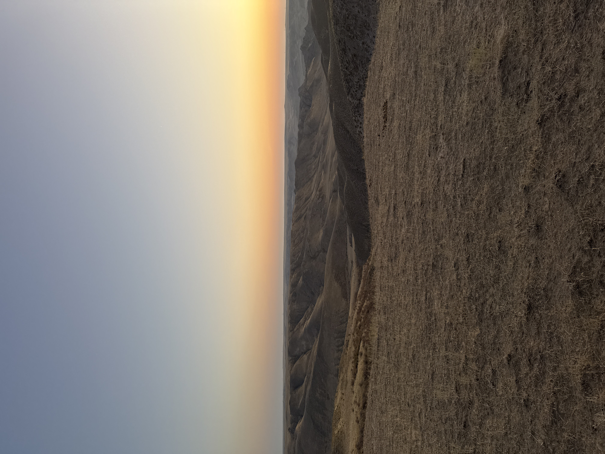 The image depicts a vast, rolling landscape with undulating hills and valleys, likely at sunset or sunrise. The sky transitions from a pale blue at the top to a warm orange and yellow gradient near the horizon, suggesting twilight. The terrain in the foreground and midground is covered in dry, brown grass and sparse vegetation, indicative of a semi-arid climate. The distant horizon shows a line of hills and possibly some faint man-made structures. The scene is natural and appears uninhabited, with no people or animals visible. The location is identified as the city of Qonaev, Kazakhstan, and the landscape suggests a remote, undeveloped area surrounding it.