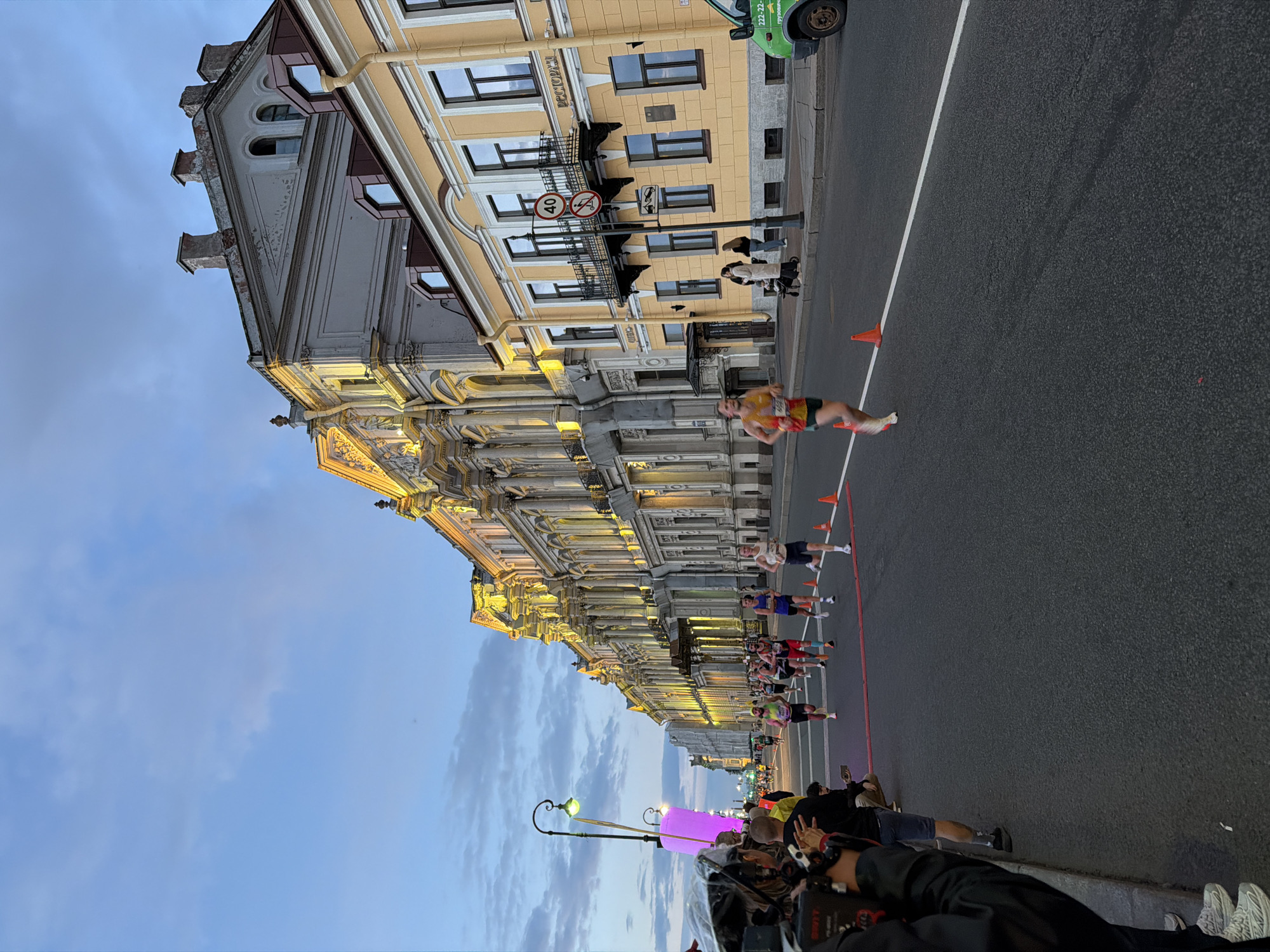 The image shows a marathon race at dusk in Saint Petersburg, Russia, likely on Nevsky Prospekt, given the grand, illuminated architecture characteristic of the city's main avenue. A group of male runners are nearing a finish line marked with red tape across the asphalt street. The buildings lining the street are ornate and historic, with yellow and gray facades, illuminated by warm lighting. Some runners are wearing bib numbers, indicating they are participants in a timed event. Spectators are visible on the left side of the road, including individuals holding cameras and one person crouching. Traffic cones are placed along the course. The sky is a pale blue with some clouds, suggesting it's early evening. A street lamp with a distinctive green light is also visible, a common feature in Saint Petersburg. A green utility vehicle is parked on the right side of the street.
