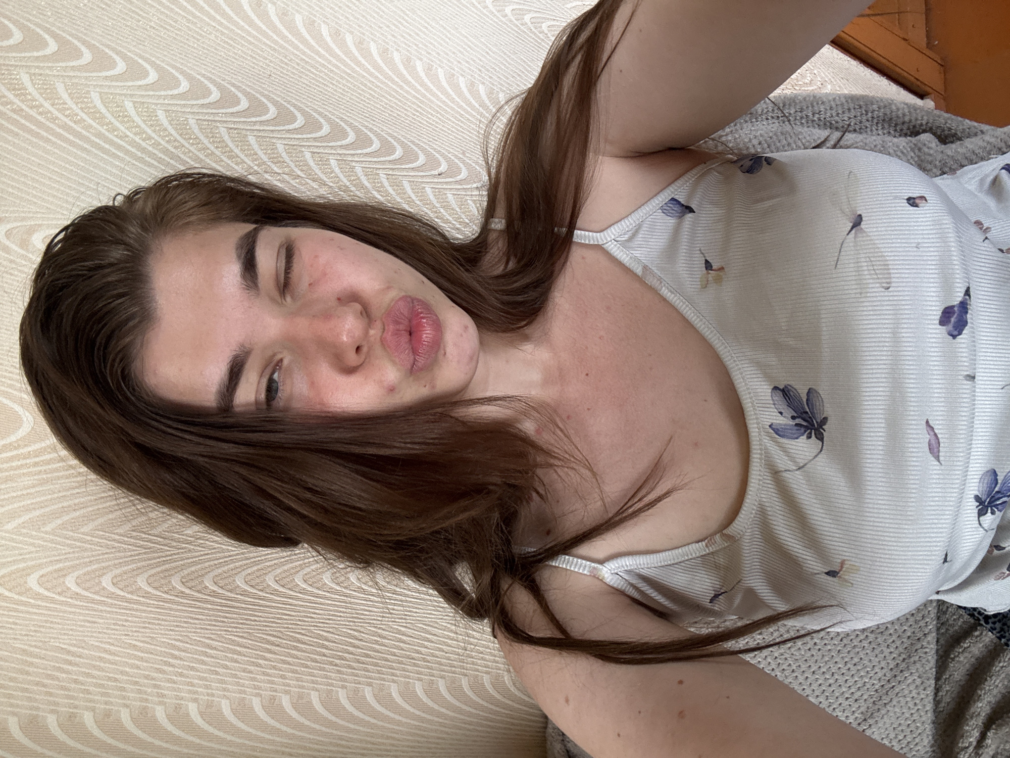 A young woman with long, brown hair is pictured from the chest up, winking and puckering her lips. She is wearing a light-colored, ribbed tank top with a floral pattern. The background is a beige, textured wallpaper with a wave-like design. The woman appears to be indoors, possibly in a residential setting. There is no visible indication of the city of Мещёры, Russia.