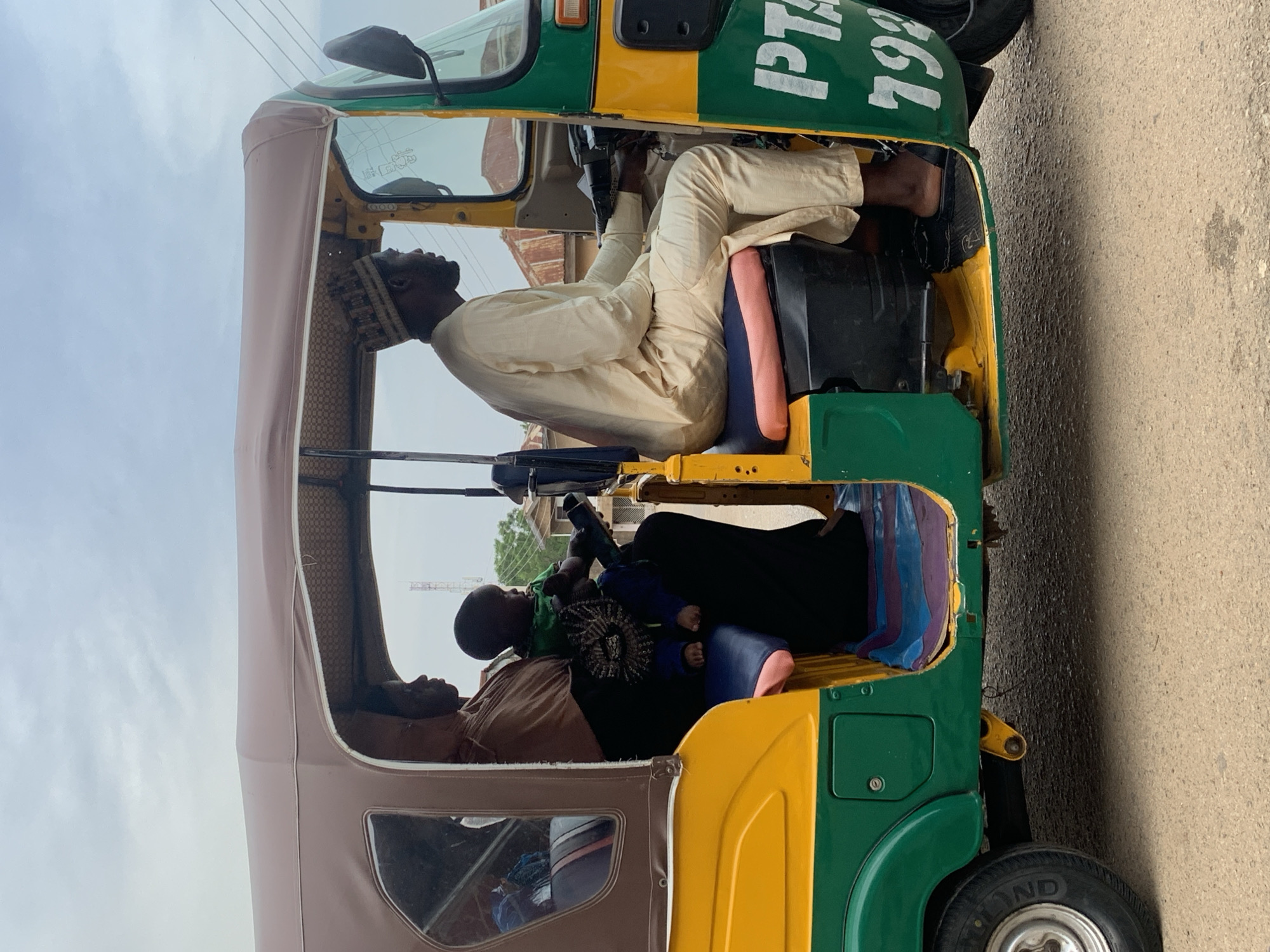 A motorized tricycle, painted yellow and green, is parked on a paved street. The driver, a dark-skinned male wearing a cream-colored robe and a kufi cap, is seated inside. Two passengers are in the back: a dark-skinned woman, and a baby lying on her lap. The registration number "PTA 193" is painted in white on the tricycle's front. The location is Jos, Nigeria.