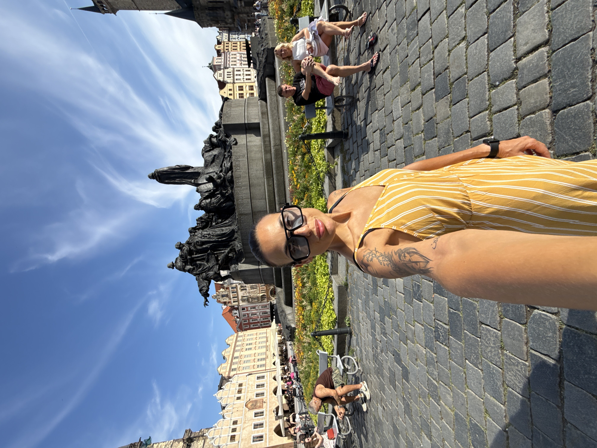A woman is taking a selfie in the Old Town Square in Prague, Czechia, on a sunny day. She is wearing a yellow and white striped dress and large black sunglasses. Behind her, a large bronze statue stands in front of a bed of colorful flowers. In the background, several historic buildings with ornate architecture line the square. To the right and left of the statue, people are seated on benches, some looking at the statue, others engaged in conversation. The ground is paved with cobblestones. The sky is blue with wispy white clouds.