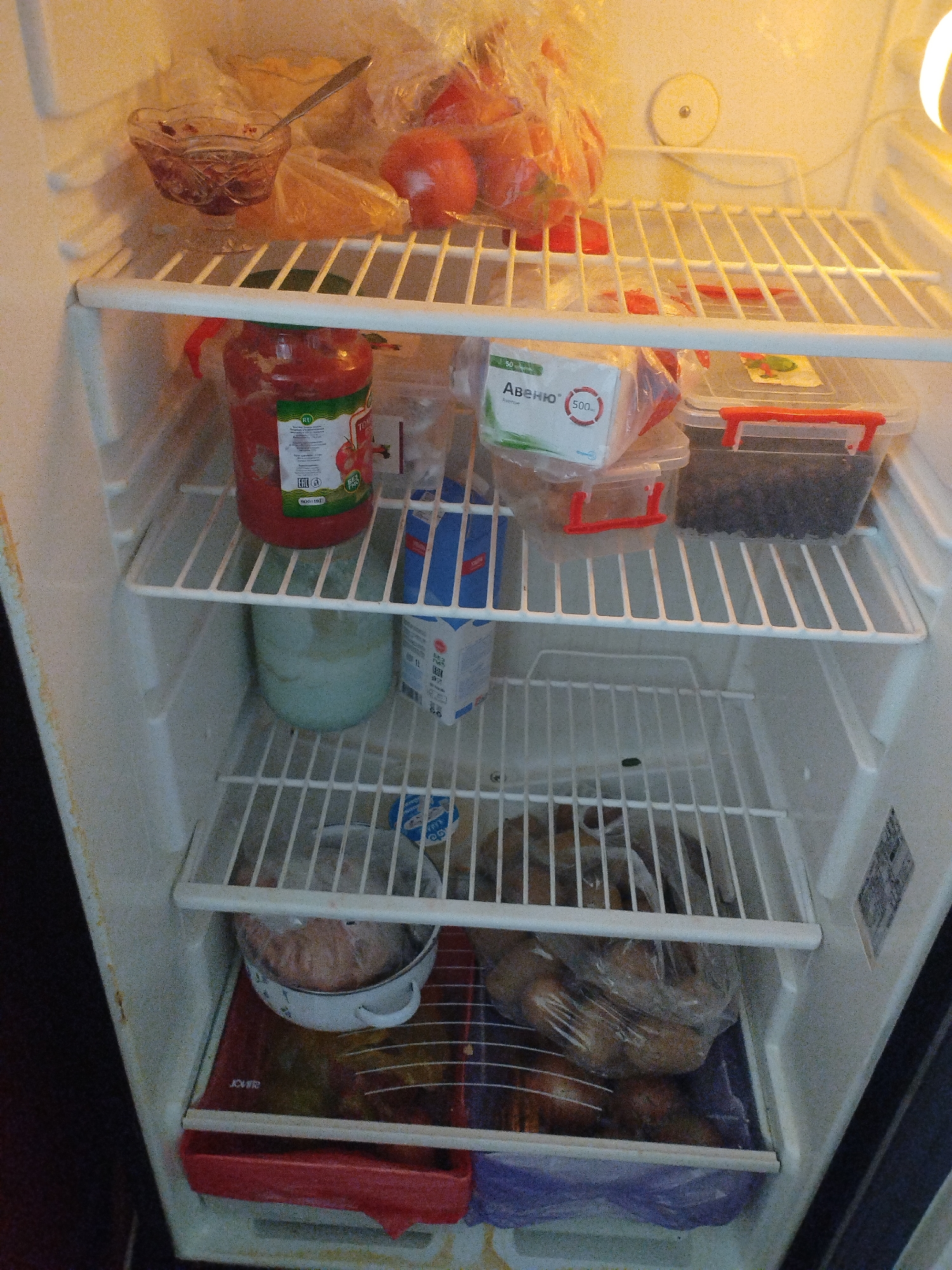 The image depicts the interior of a refrigerator, stocked with various food items. On the top shelf, there is a glass bowl containing a dark red substance, possibly jam or preserve, with a spoon resting in it. Behind the bowl, a plastic bag contains what appear to be tomatoes. To the right of the tomatoes, another plastic bag is visible.
The second shelf from the top holds a large red jar of tomato paste or sauce, identifiable by the "RU" label and a graphic of tomatoes. Next to it, a white carton with blue printing, possibly milk or juice, is present. Also on this shelf is a transparent plastic container with a red lid, containing a dark, granular substance, likely berries. Another similar container, with a red lid, sits beside it.
The third shelf from the top contains a light blue, opaque container, likely dairy-based, such as yogurt or kefir. There is also a small blue and white packet with text in a foreign language.
The bottom shelf displays a white bowl with handles, containing a pinkish-brown substance, possibly raw meat or a meat mixture. This bowl rests on a red tray with some markings. Beneath the tray, on the floor of the refrigerator, is a purple plastic bag containing more food items. The overall environment of the refrigerator appears to be standard for a home appliance.
The location is specified as the city of Октябрь, Kyrgyzstan, although this information cannot be visually verified from the image alone.