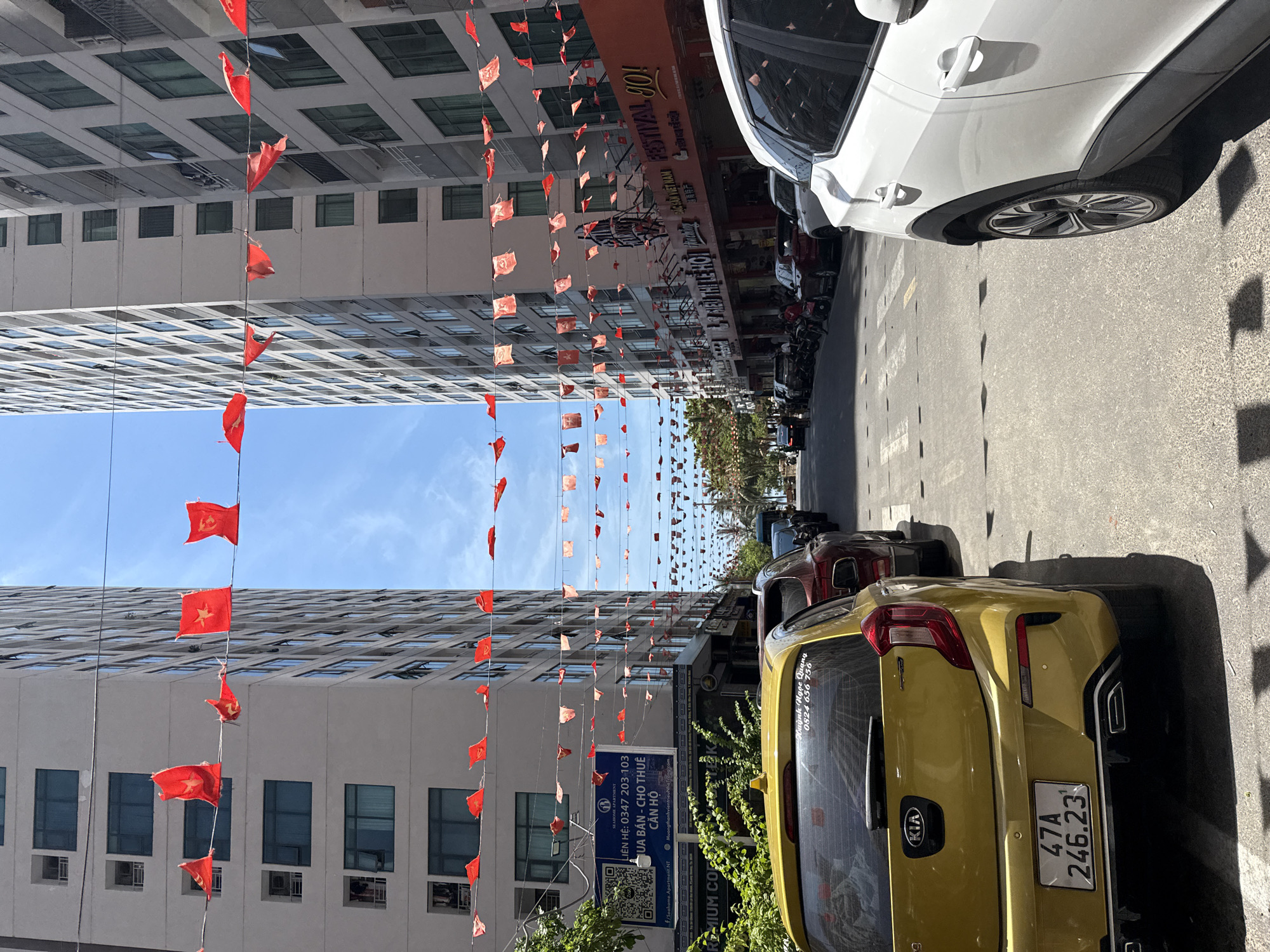 The image depicts a street scene in Nha Trang, Vietnam, characterized by tall, modern buildings lining both sides of the road. Red and yellow flags, many appearing tattered, are strung across the street at various heights, creating a festive or commemorative atmosphere. The road is paved and marked with white lines, and several cars are parked or driving on it, including a yellow Kia in the foreground. There are also parked motorcycles visible. The architecture of the buildings suggests a commercial or residential urban environment. The sky is blue and clear, indicating fair weather. The overall impression is one of a busy street in a Vietnamese city, decorated for an event or holiday.